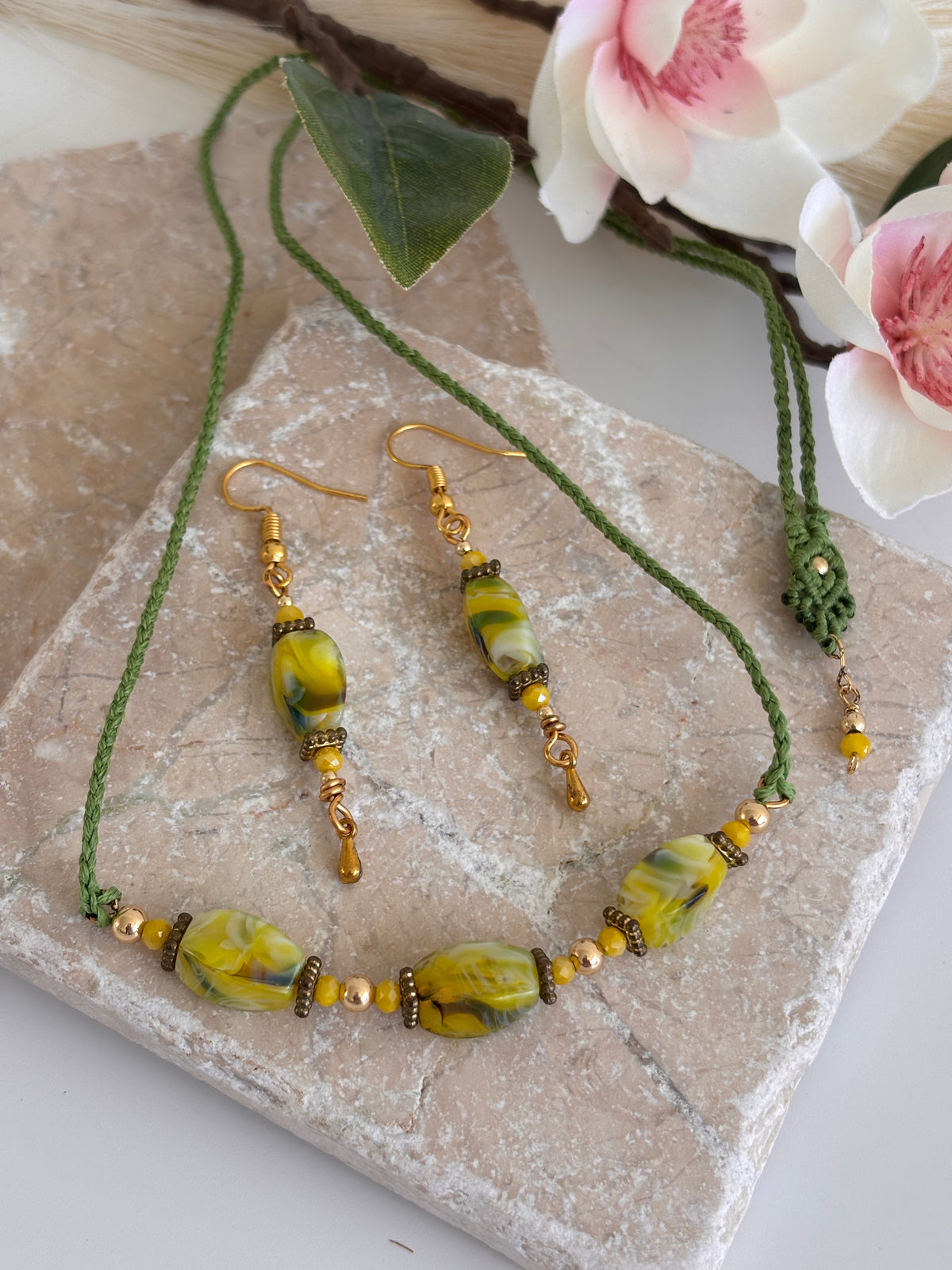 Handmade chartreuse twisted rectangle lampwork glass drop earrings with square gold-tone spacer beads, 3mm opaque yellow crystal, and 3mm gold-tone teardrop dangle shown styled with coordinating green macrame lampwork glass necklace on travertine tile with white magnolia flowers.