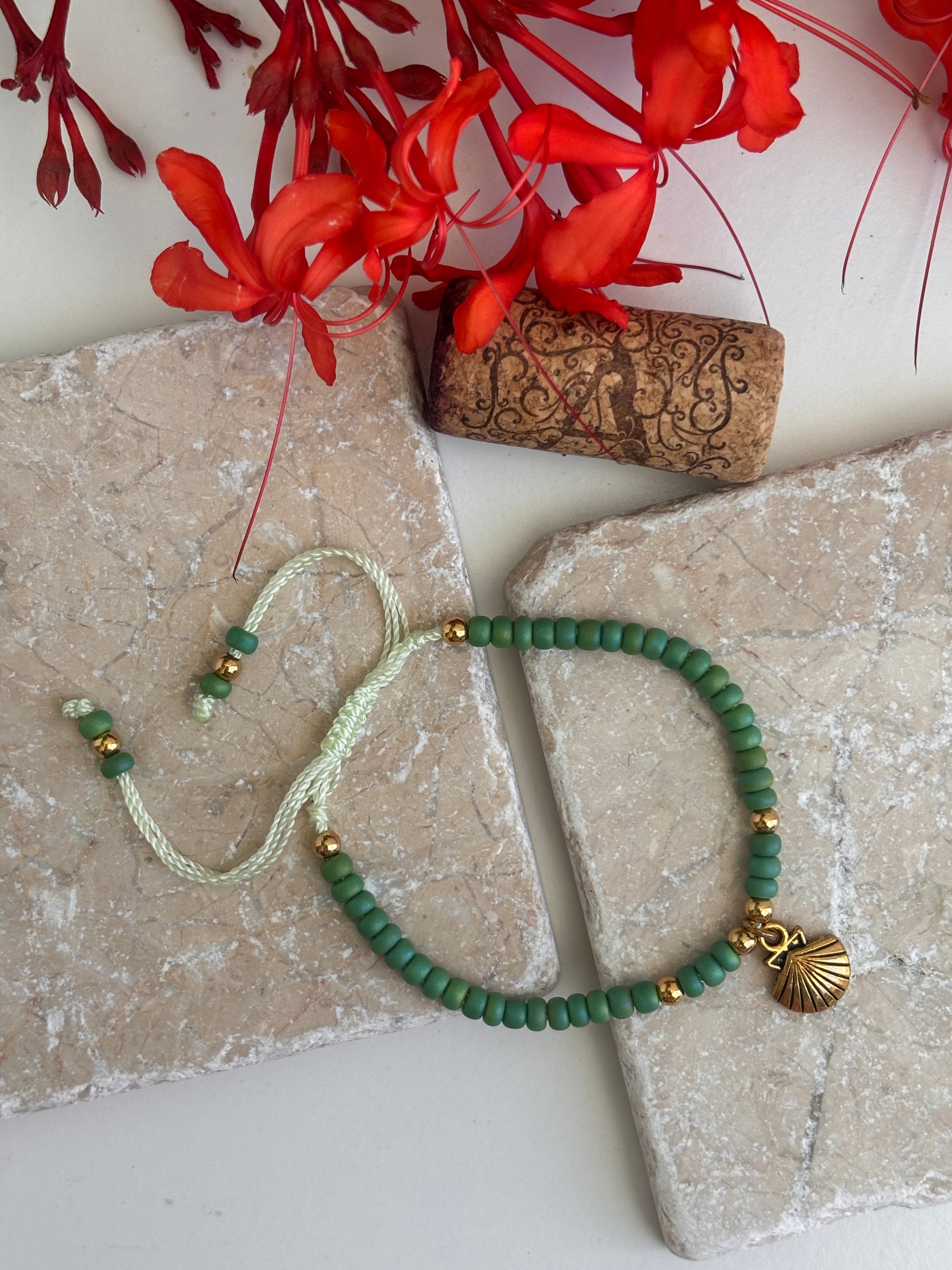 A beaded bracelet with 6/0 green round beads, a mint green adjustable cord, and a brass shell charm, styled on stone tiles with red flowers and a cork in the background.