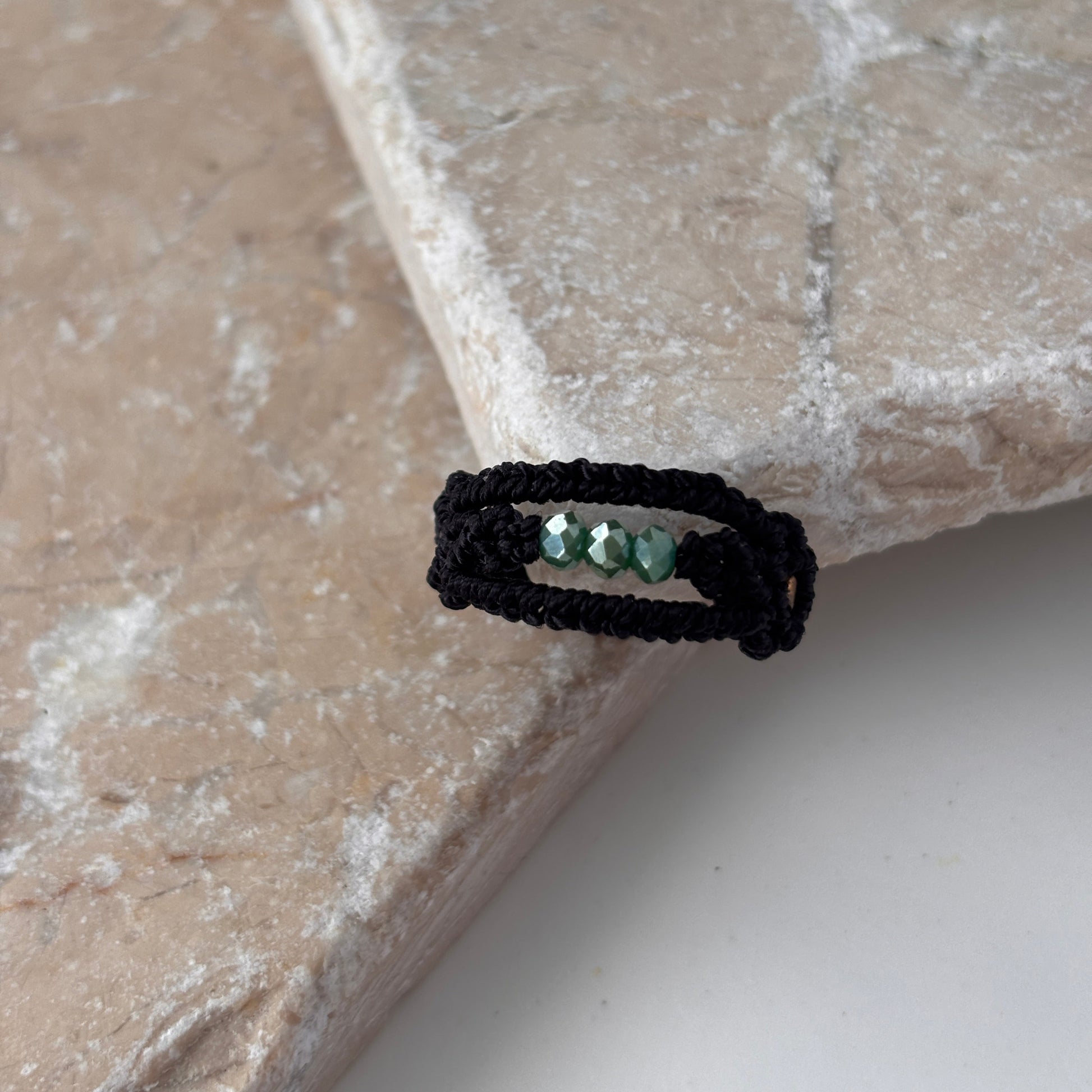 A top-down view of a handmade macrame band ring in black flat waxed nylon cord with a double-wrap braided design and three faceted mint green glass crystal beads across the center face, resting on the edge of a pink stone surface against a white background.