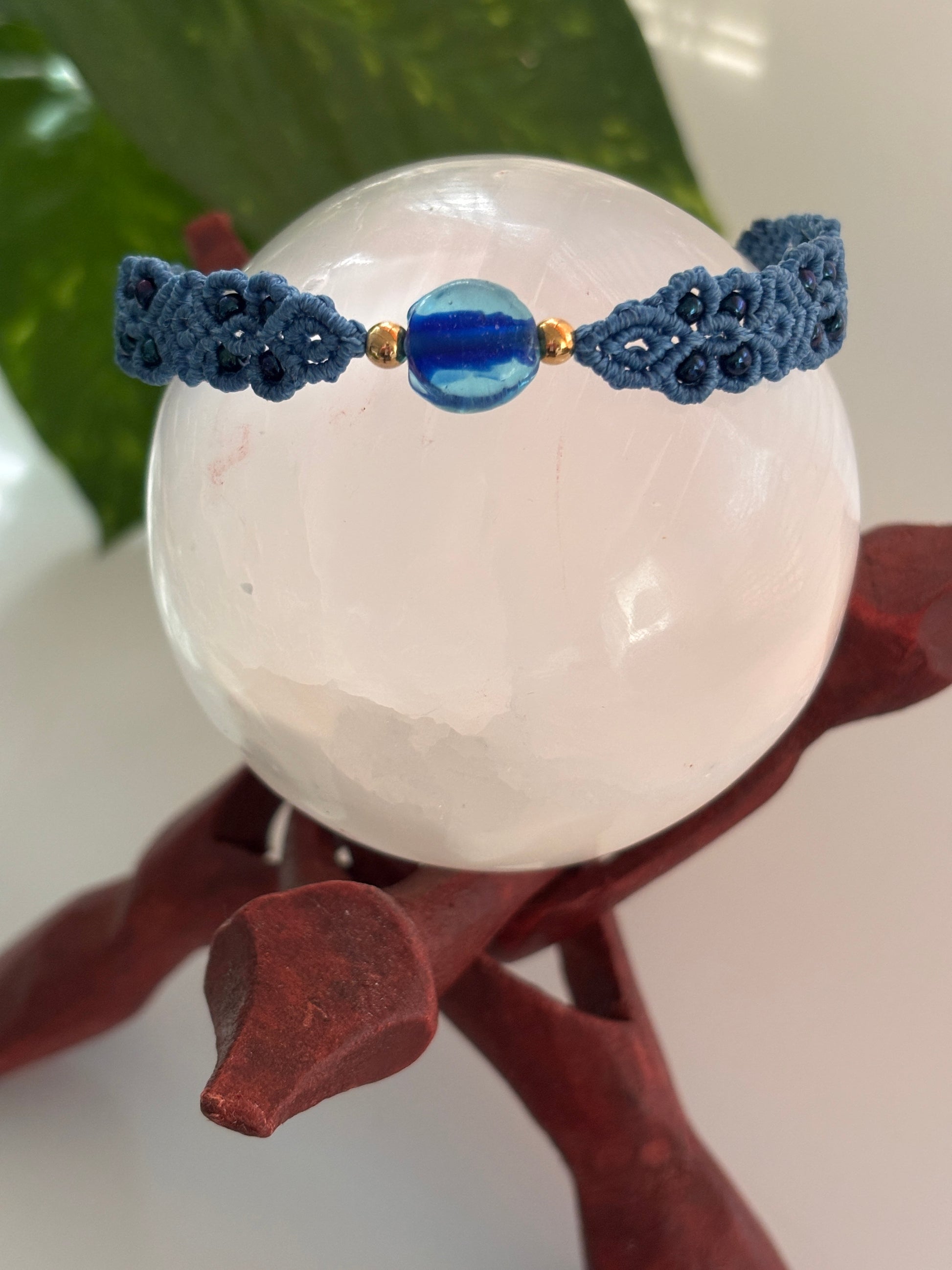 A medium blue macrame bracelet with a central aqua blue glass disc bead and small brass spacer beads, displayed on a white crystal sphere stand.