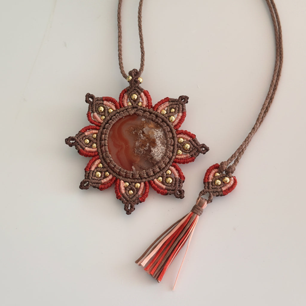 A handmade macrame pendant necklace featuring a central Carnelian Agate cabochon stone surrounded by rust, peach, and brown woven sunflower mandala medallion with small brass beads, shown on a neutral light gray background with a matching tassel detail.
