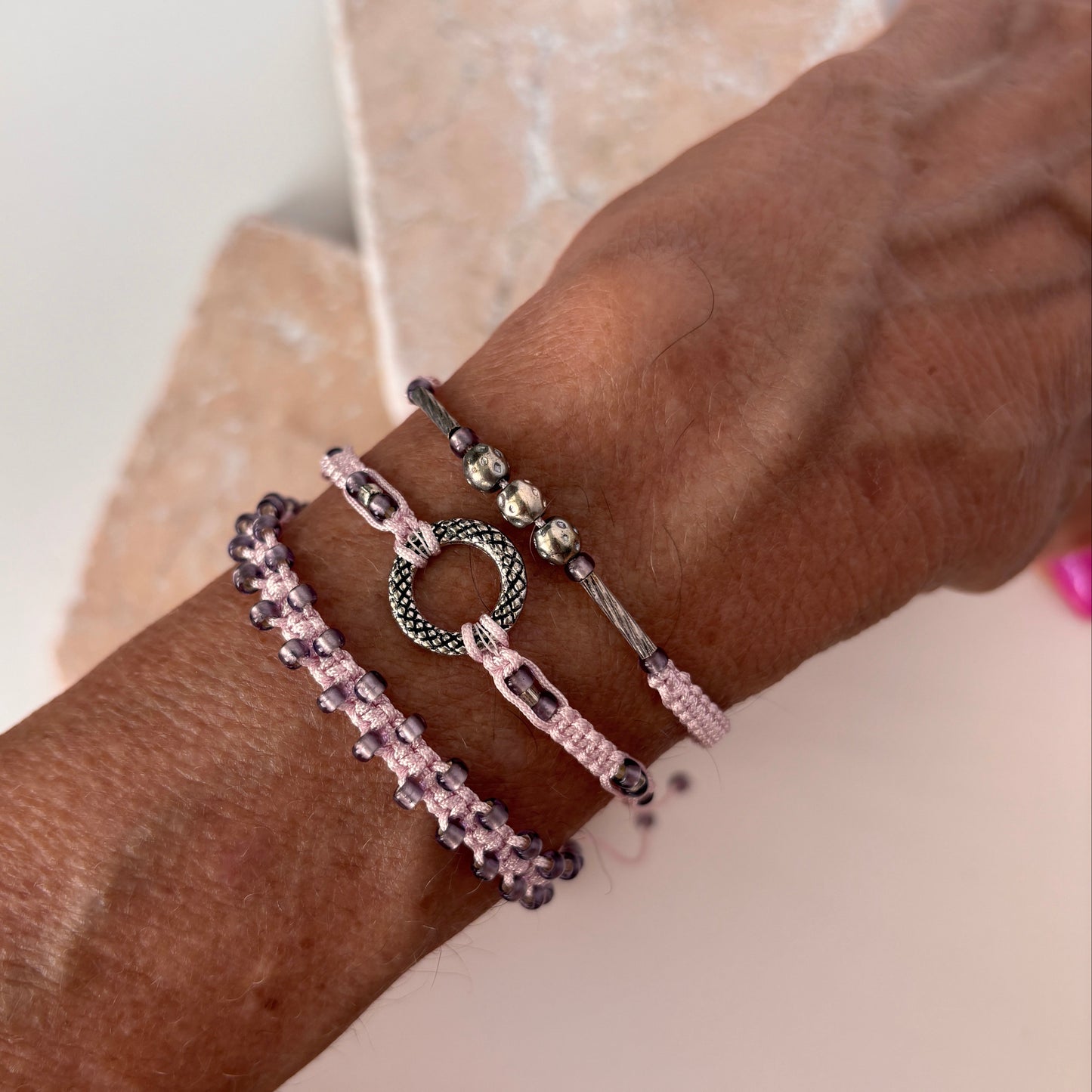 A close-up of three light purple macrame bracelets stacked on a wrist against a pink stone surface — one with lavender seed beads woven along the edges, one with a silver-tone open ring centerpiece, and one with a silver-tone tube and silver-tone ball accent beads on light purple nylon cord.