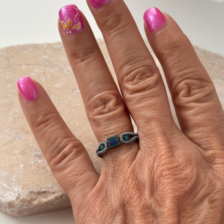 A front view of a handmade minimalist gray macrame ring with an iridescent blue-purple square Miyuki Tila bead at the center and two teal seed bead accents, worn on a finger resting on a pink stone surface with pink nail art in the background.