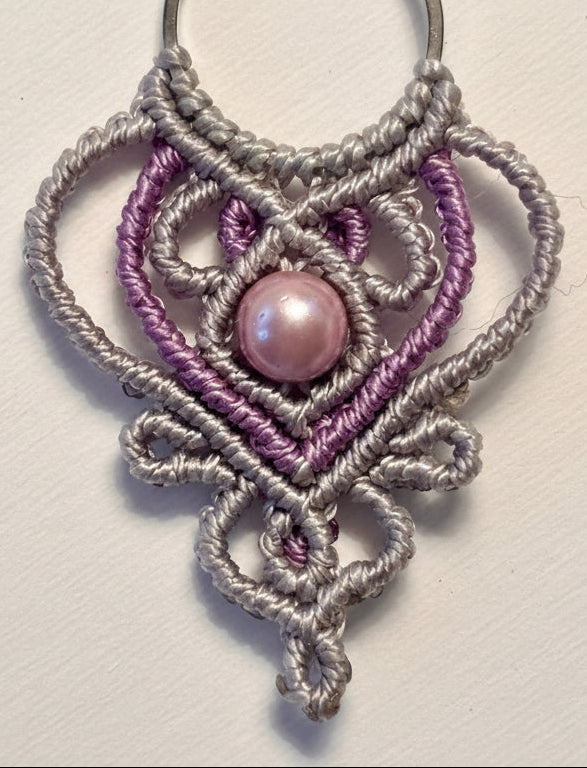 Macro close-up of handmade two-tone silvery gray and light purple macrame teardrop drop earring showing ornate heart-shaped lattice knotwork detail and 6mm mauve shell pearl center bead.