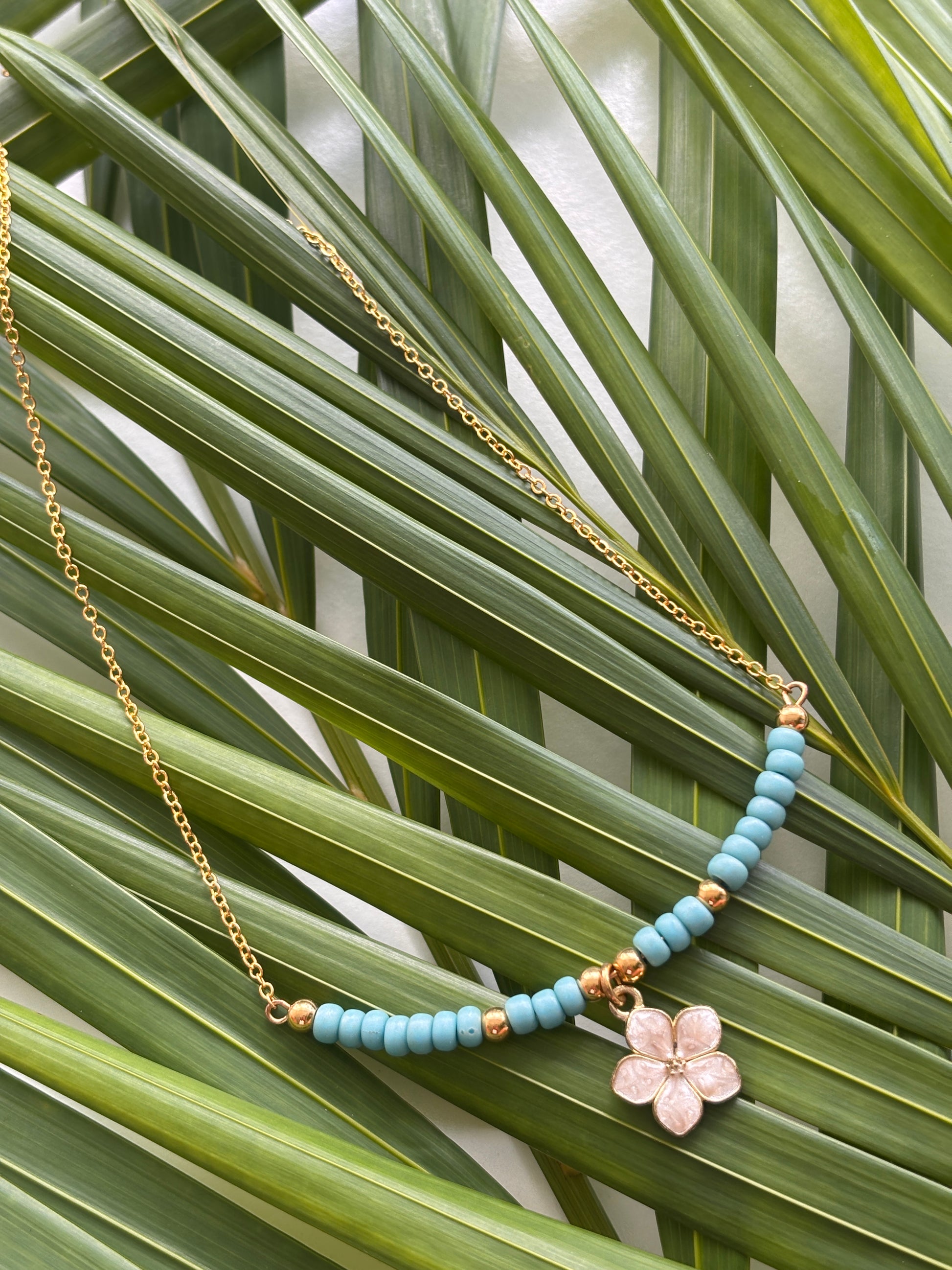 A brass chain necklace with turquoise blue seed beads, small brass spacer beads, and a white enamel flower charm, displayed on green palm leaves.