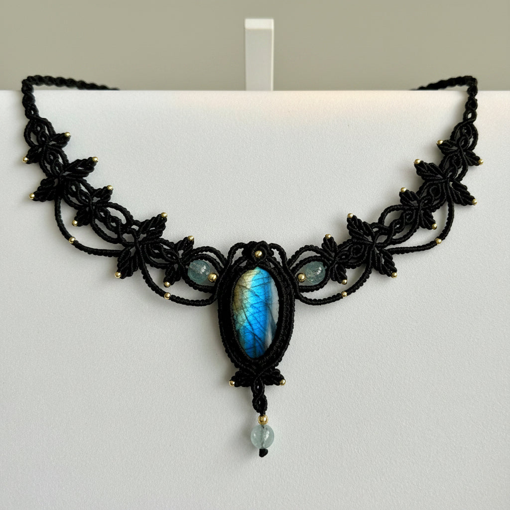 Black macrame-style necklace with a central oval blue-gray stone and small light blue beads, photographed on a plain light background.