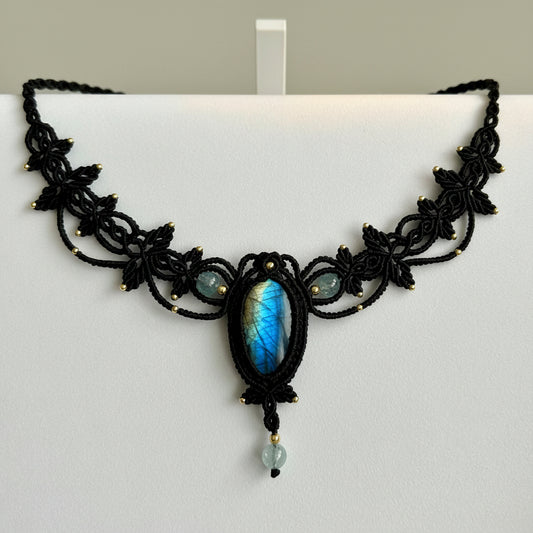 Black macrame-style necklace with a central oval blue-gray stone and small light blue beads, photographed on a plain light background.