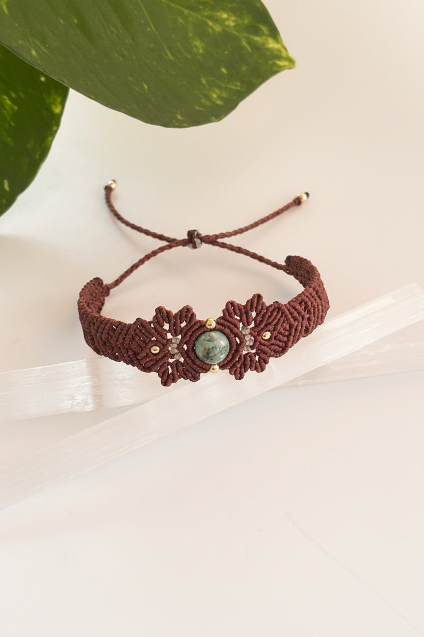 A brown macrame bracelet with a central Indian Agate green stone and small brass bead accents, displayed on a white surface with a green leaf in the background.