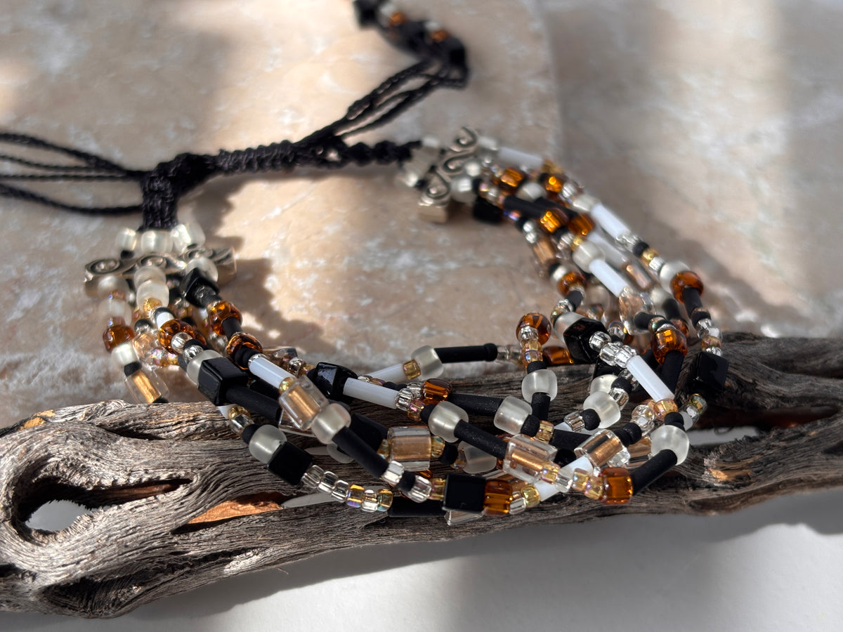 A close-up of a handmade seven strand seed bead bracelet in black nylon macrame cord with mixed frosted white, matte black, amber rootbeer, and clear glass seed beads, cube beads, and bugle beads with silver-tone scroll spacer bars, draped over weathered driftwood against a pink stone background in natural sunlight.