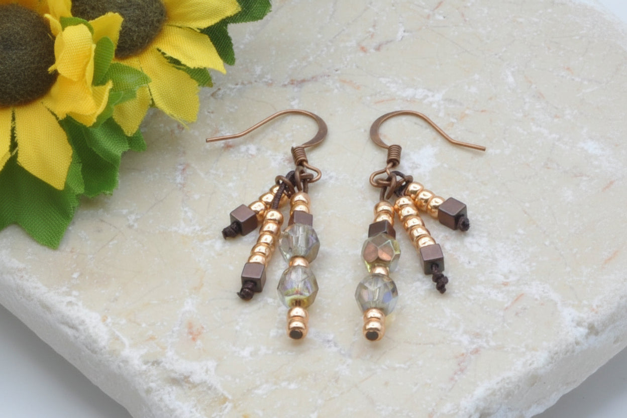 Handmade beaded drop earrings with copper wires, gold seed beads, crystal and cube beads displayed on stone with sunflower — boho jewelry by MJ Skivers Designs.