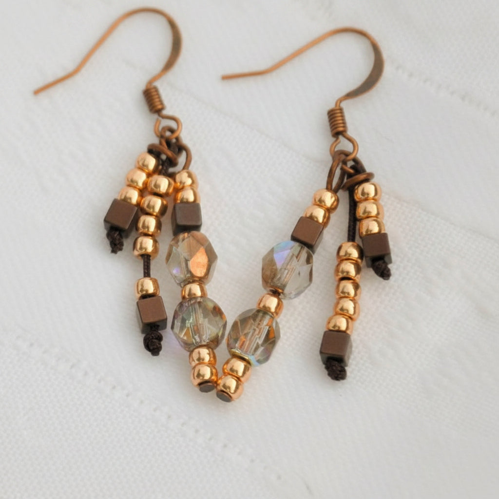 Handmade beaded drop earrings with three strands of gold seed beads, dark hematite cube beads, and iridescent faceted crystal beads on copper-tone fish hook wires, displayed on a white surface.