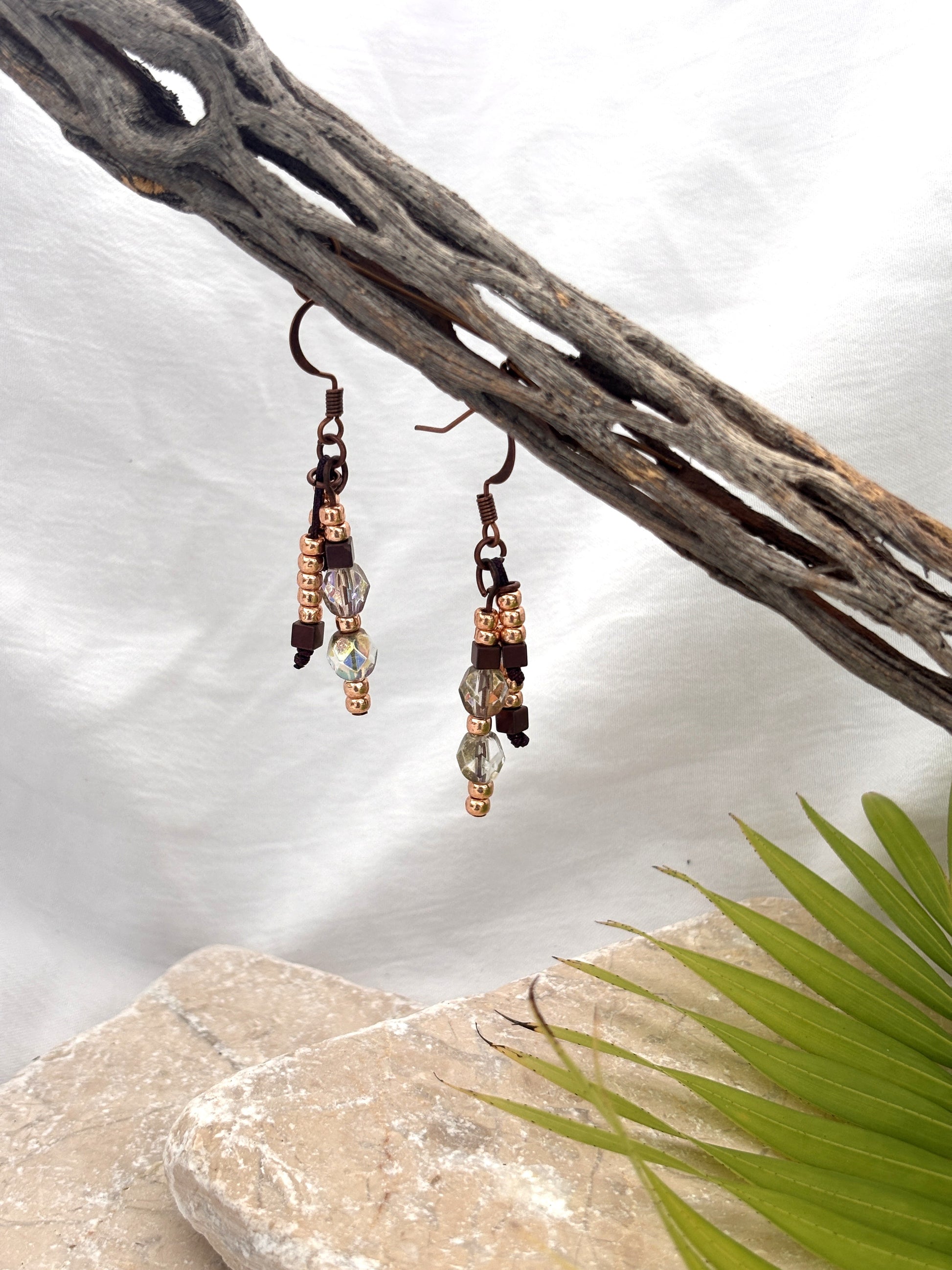 Handmade beaded drop earrings with gold seed beads, hematite cube beads, and iridescent faceted crystal beads on copper-tone fish hook wires, hanging from a driftwood branch with a stone and palm leaf backdrop.