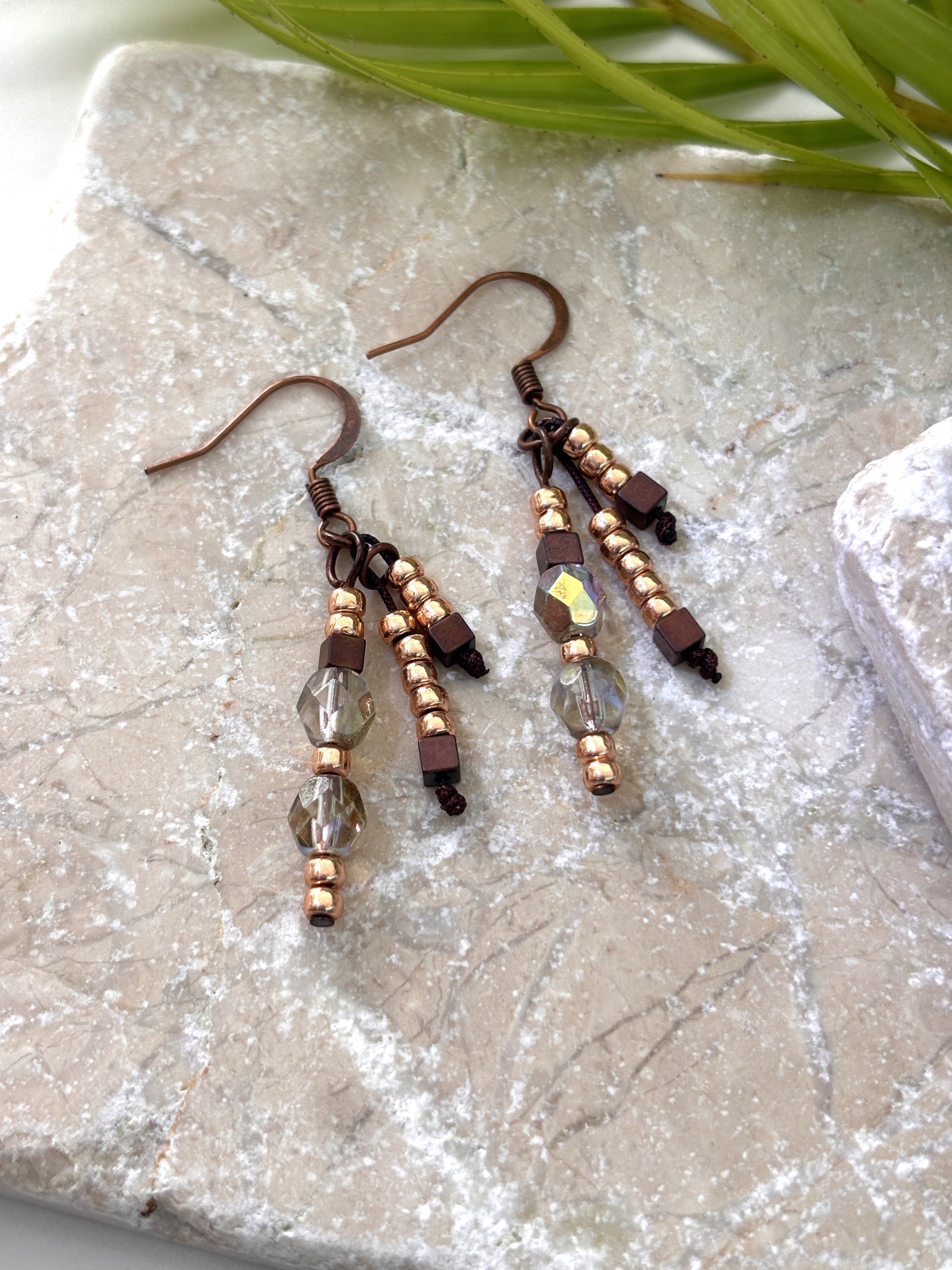 Pair of handmade beaded drop earrings with gold seed beads, hematite cube beads, and iridescent faceted crystal beads on copper-tone fish hook wires, laid flat on a natural stone surface with a palm leaf.