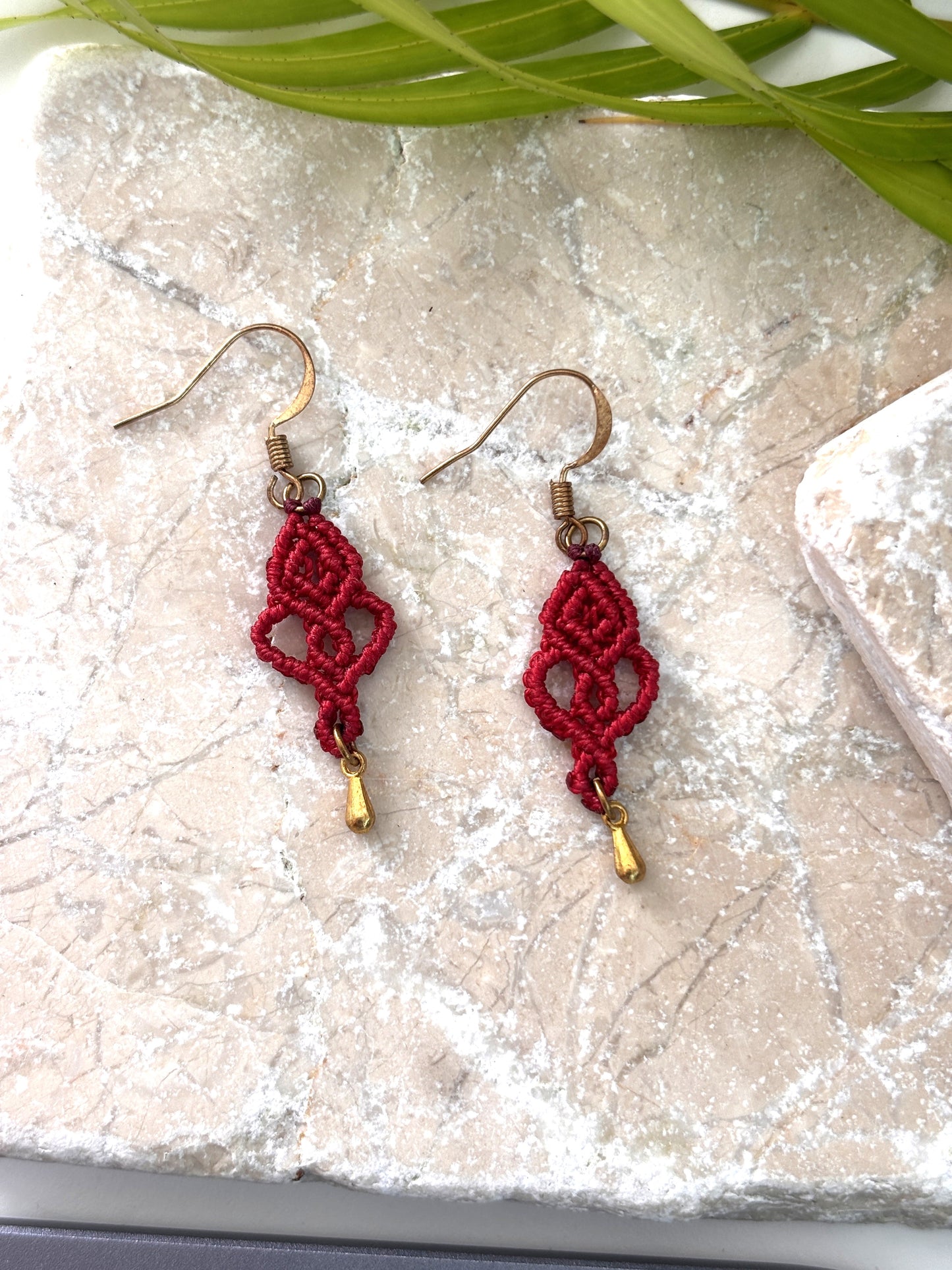 Pair of handmade red macrame looping hearts dangle earrings with gold-tone teardrop charms and gold-tone fish hook wires, laid flat on a natural stone surface with a palm leaf.