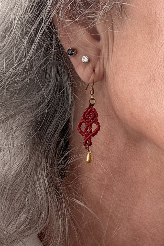 Close-up of a handmade red macrame looping hearts dangle earring with a gold-tone teardrop charm and gold-tone fish hook wire, worn on a model's ear.