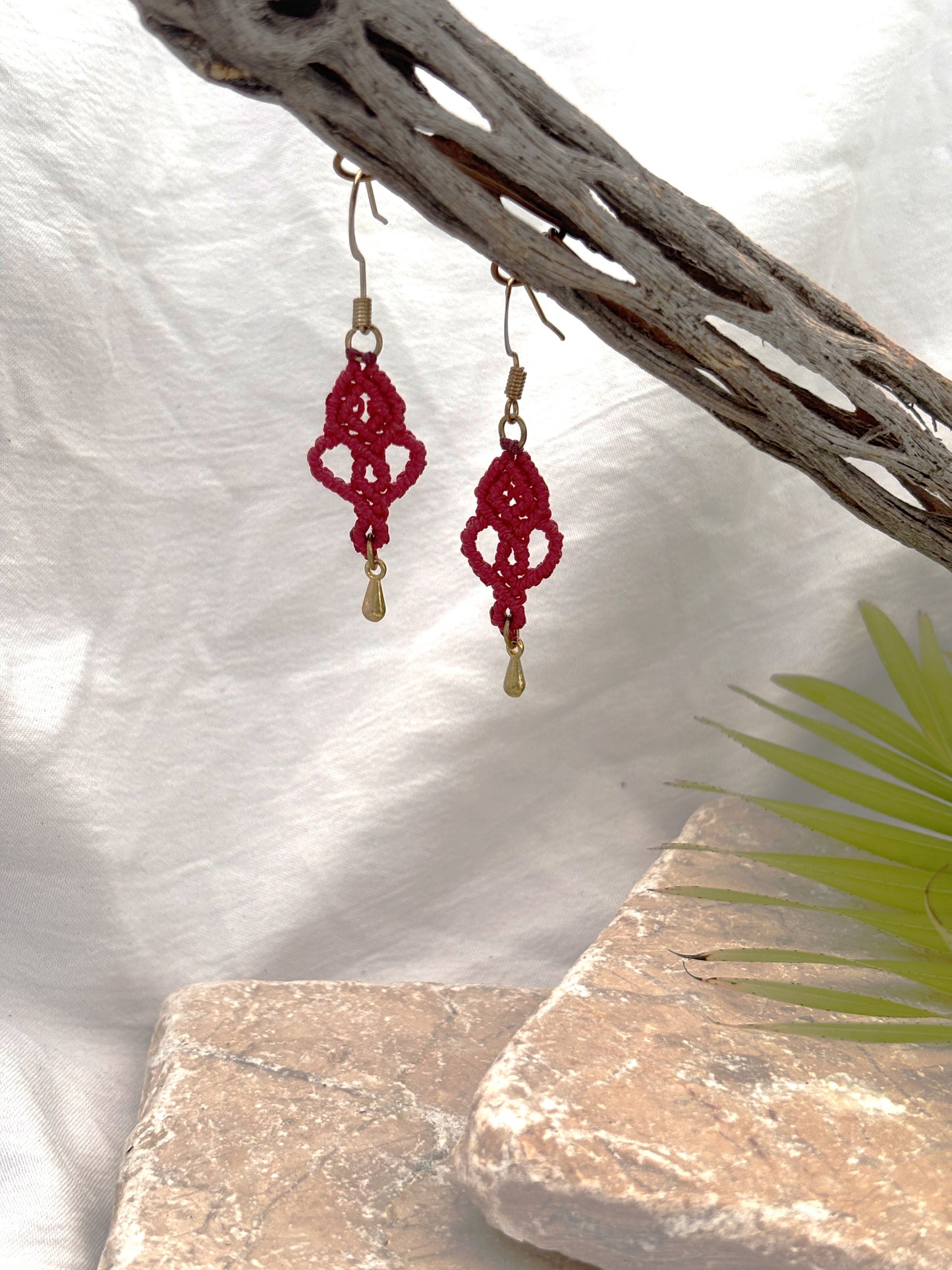 Handmade red macrame looping hearts dangle earrings with gold-tone teardrop charms hanging from a driftwood branch with a stone and palm leaf backdrop.