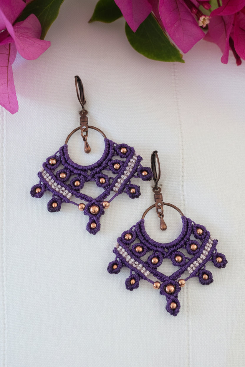 A pair of handcrafted macrame chandelier earrings in deep purple nylon cord with a fan shape, open central ring, copper round beads woven throughout, a lavender macrame cord accent row across the center, and antique copper lever back ear wires with a small copper teardrop dangle, displayed on a white surface with vibrant magenta bougainvillea flowers and green leaves in the background.