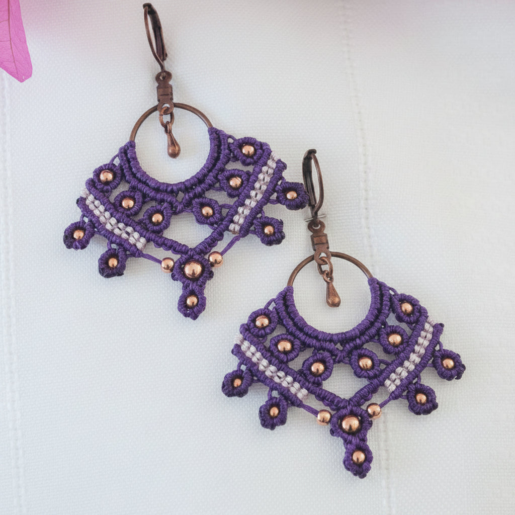  close-up of a pair of handcrafted macrame chandelier earrings in deep purple nylon cord with a fan shape, open central ring, copper round beads woven throughout, a lavender macrame cord accent row across the center, and antique copper lever back ear wires with a small copper teardrop dangle, displayed on a white surface with a soft-focus pink flower in the background.