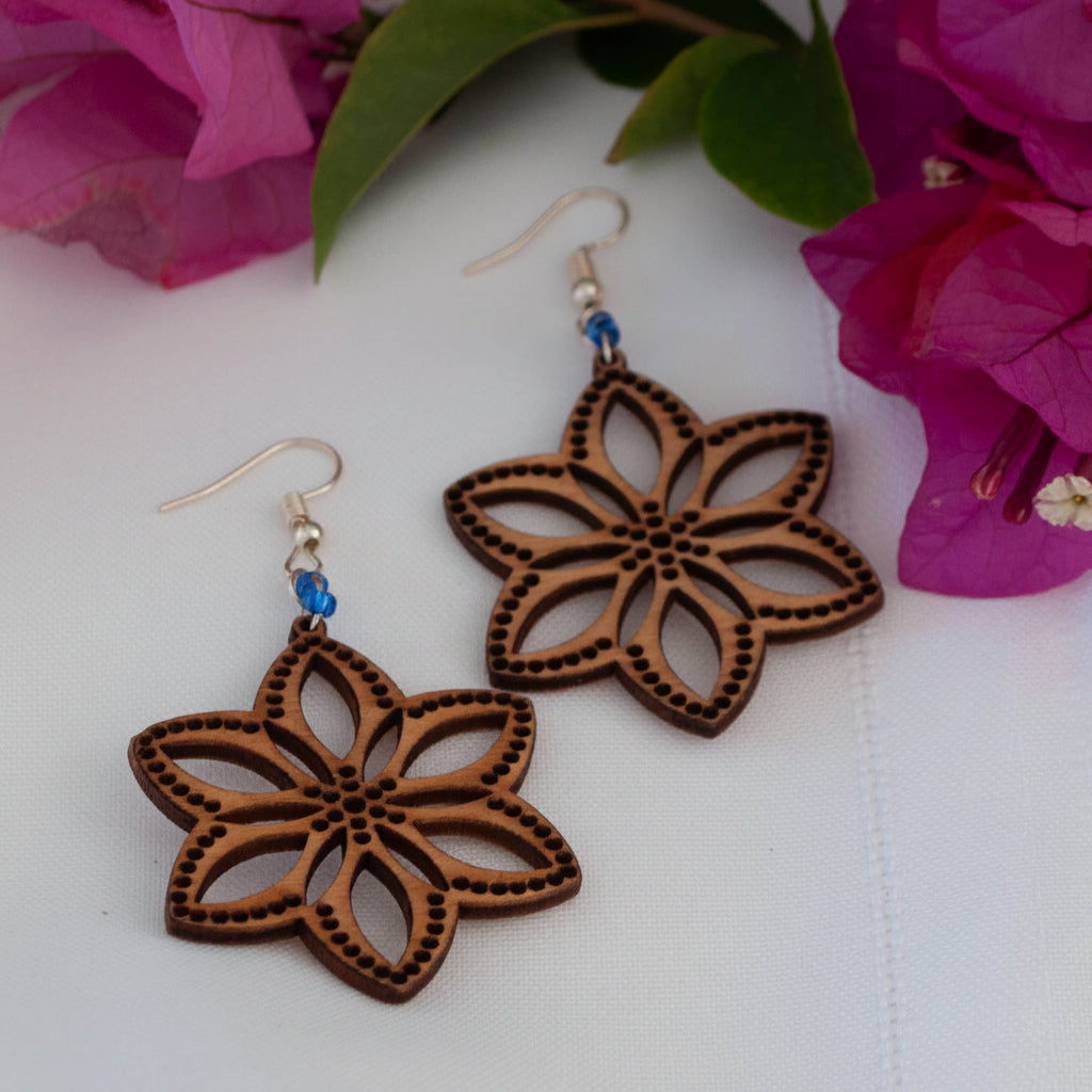 Handmade balsa wood flower earrings with cornflower blue seed beads and silver-tone fish hook wires, displayed on a white surface with magenta bougainvillea flowers.