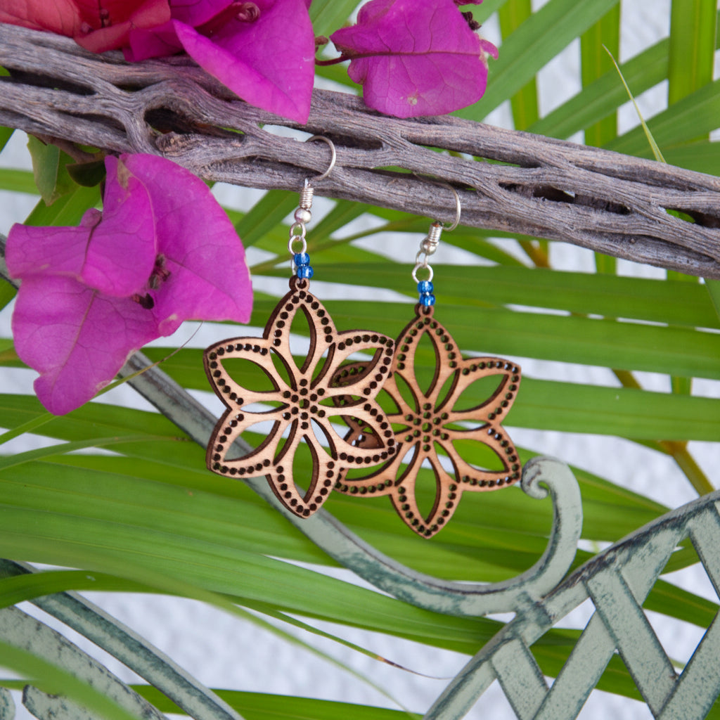 Handmade balsa wood flower earrings with cornflower blue seed beads and silver-tone fish hook wires, hanging from a driftwood branch with tropical palm leaves and magenta bougainvillea flowers.