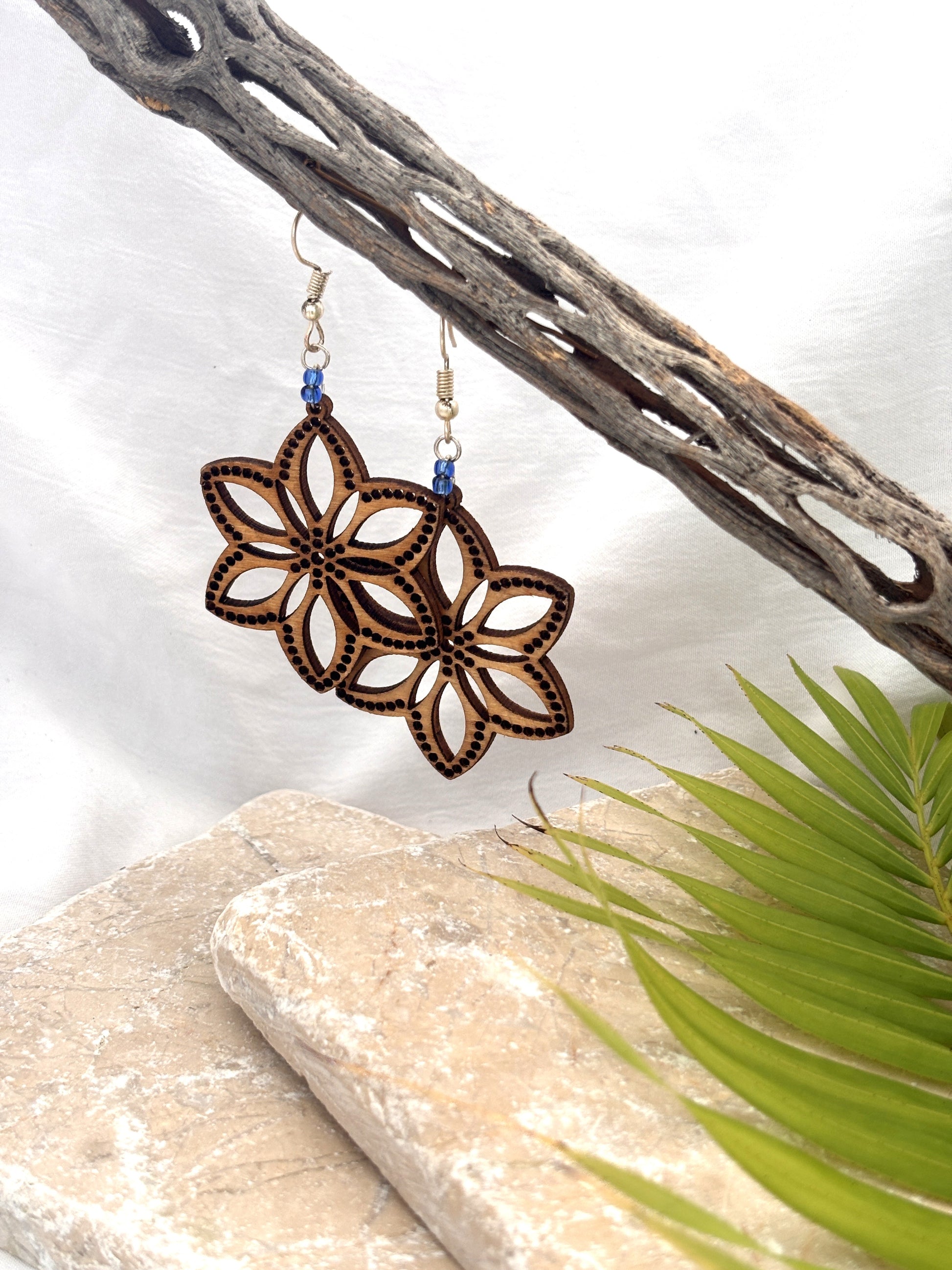Handmade balsa wood flower earrings with open petal silhouette, dotted border detail, and cornflower blue seed beads on silver-tone fish hook wires, hanging from a driftwood branch with a stone and palm leaf backdrop.
