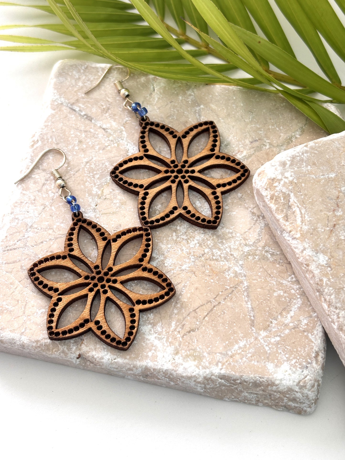 Pair of handmade balsa wood flower earrings with open petal silhouette, dotted border detail, and cornflower blue seed beads on silver-tone fish hook wires, laid flat on a natural stone surface with a tropical palm leaf.