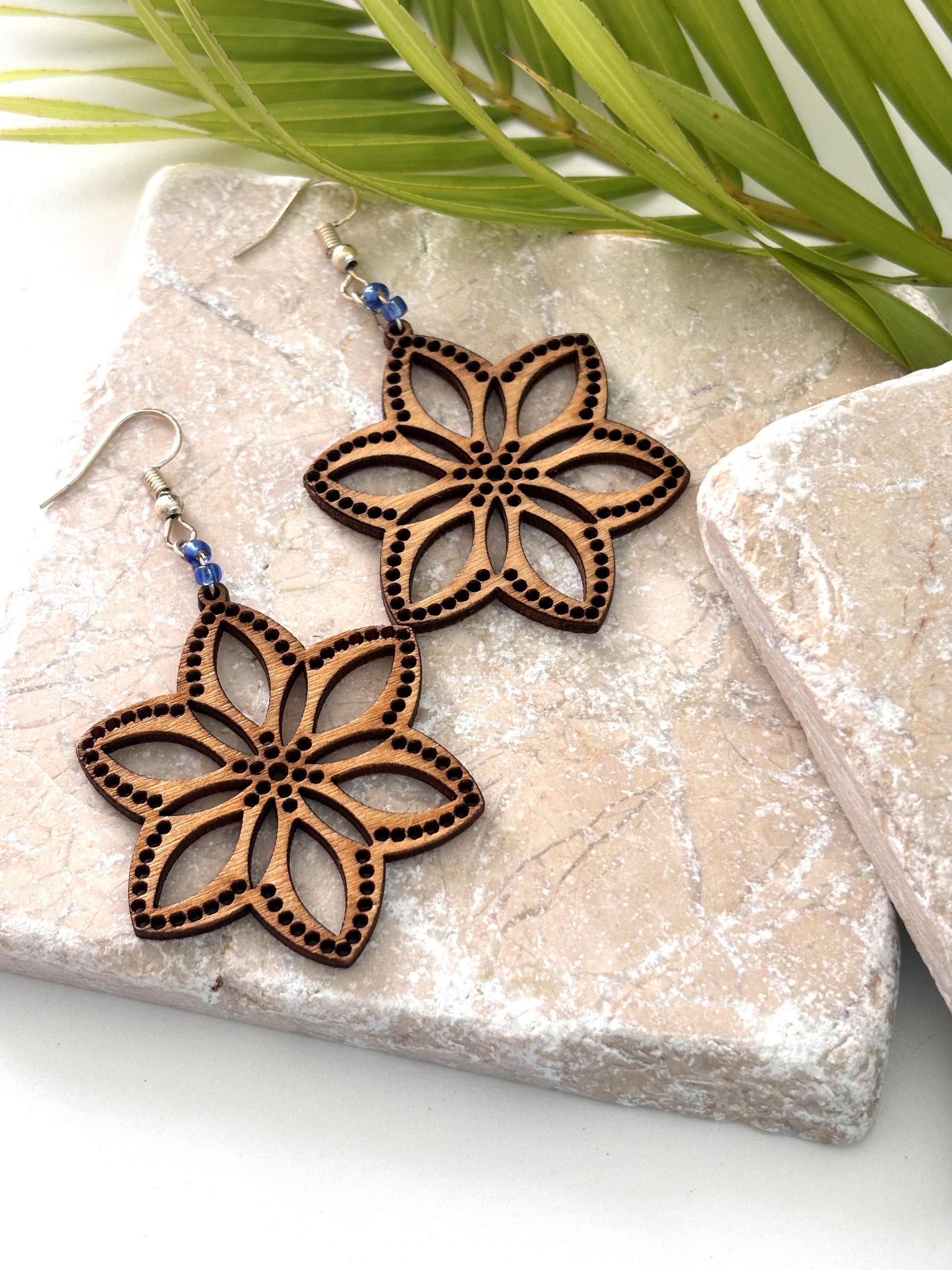Pair of handmade balsa wood flower earrings with open petal silhouette, dotted border detail, and cornflower blue seed beads on silver-tone fish hook wires, laid flat on a natural stone surface with a tropical palm leaf.