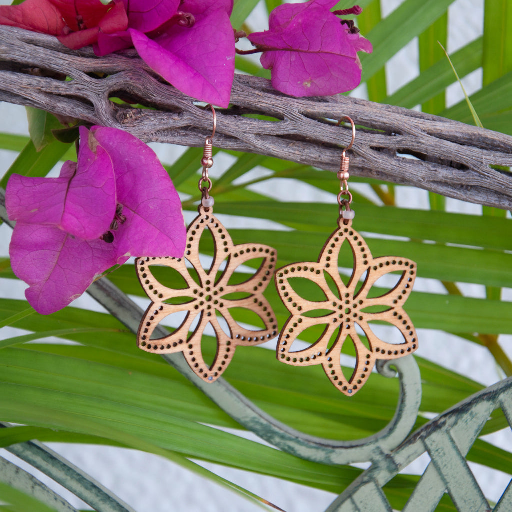 Handmade balsa wood flower earrings with open petal silhouette and dotted border detail with frosted peach seed beads on copper fish hook wires, hanging from driftwood with tropical flowers and green leaves.