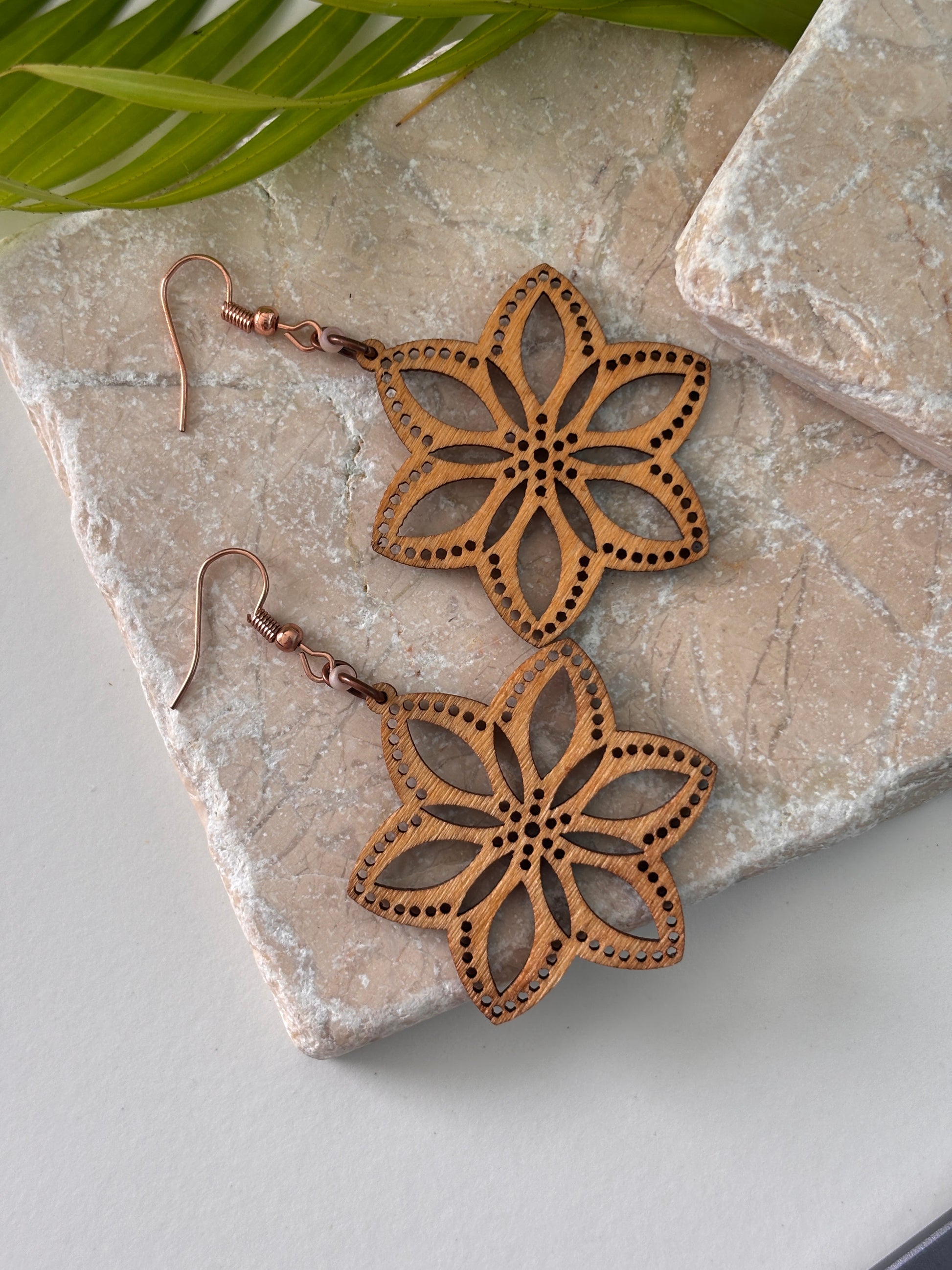 Pair of handmade balsa wood flower earrings with open petal silhouette and dotted border detail with frosted peach seed beads on copper fish hook wires, laid flat on a natural stone surface with a tropical palm leaf.