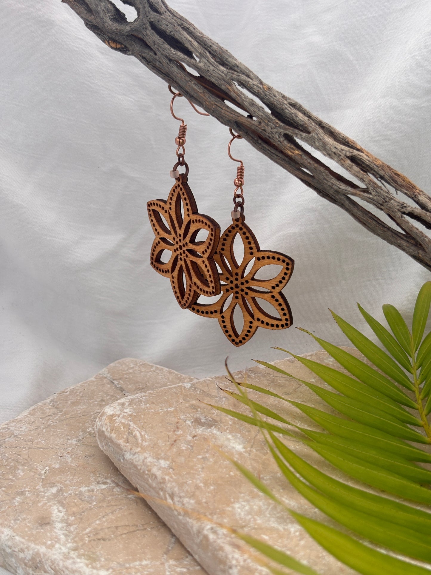 Handmade balsa wood flower earrings with open petal silhouette and dotted border detail with frosted peach seed beads hanging from a driftwood branch with a stone and palm leaf backdrop.