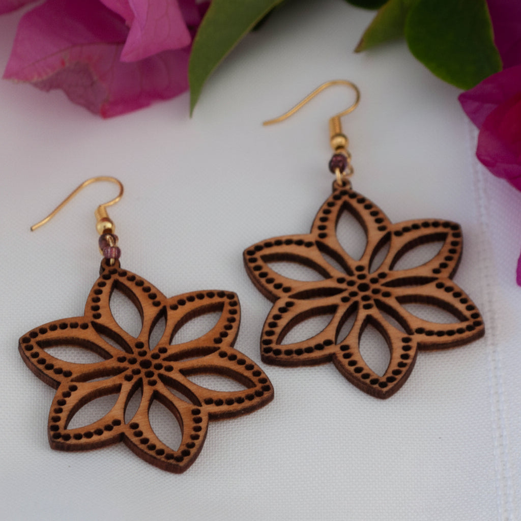 Handmade balsa wood flower earrings with plum seed beads and gold-tone fish hook wires, displayed on a white surface with magenta bougainvillea flowers in the background.