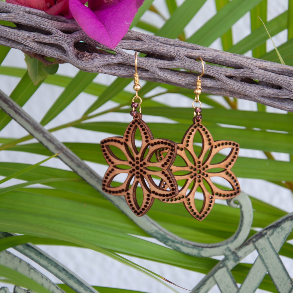 Handmade balsa wood flower earrings with plum seed beads and gold-tone fish hook wires, hanging from a driftwood branch with tropical palm leaves and magenta flowers.
