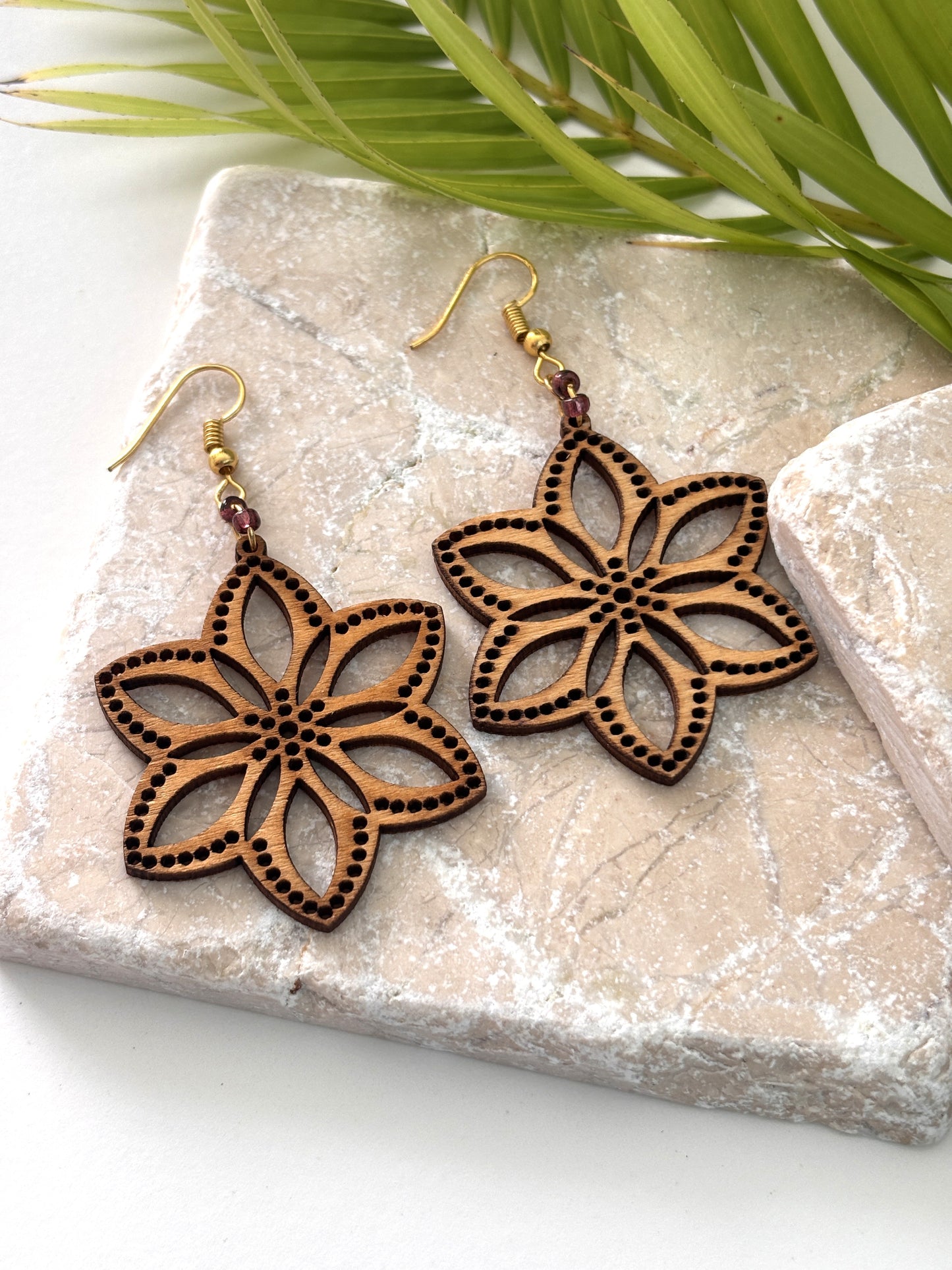 Pair of handmade balsa wood flower earrings with open petal silhouette, dotted border detail, and plum seed bead accents on gold-tone fish hook wires, laid flat on a natural stone surface with a tropical palm leaf.