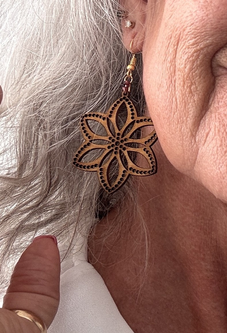 Close-up of a handmade balsa wood flower earring with an open petal silhouette, dotted border detail, and plum seed bead accent on a gold-tone fish hook wire, worn on a model's ear.