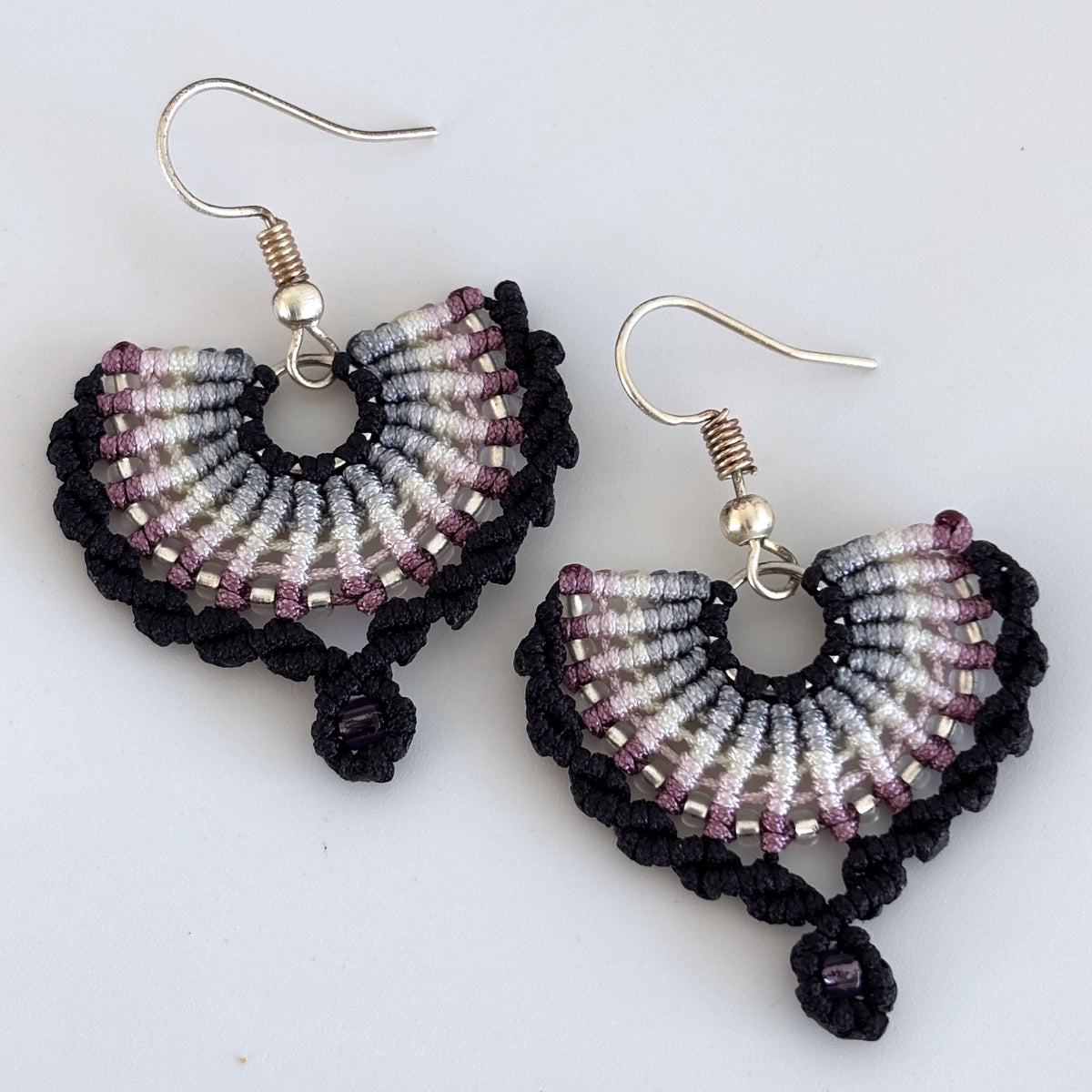 Pair of handmade black macrame fan drop earrings with semicircular fan knotwork in gradient black, steel gray, ivory, pink, and mauve cord, size 11/0 silver-lined seed bead accent rows, size 8/0 amethyst seed bead macrame loop drop, and silver-tone fish hook wires, displayed on white background showing full construction detail.