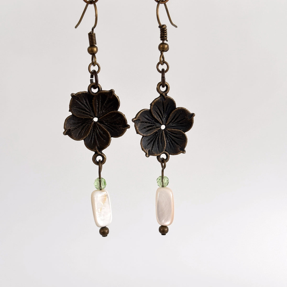 Handmade bronze flower charm earrings with mother of pearl drop, light green crystal bead, and bronze ear wires, hanging against a neutral background