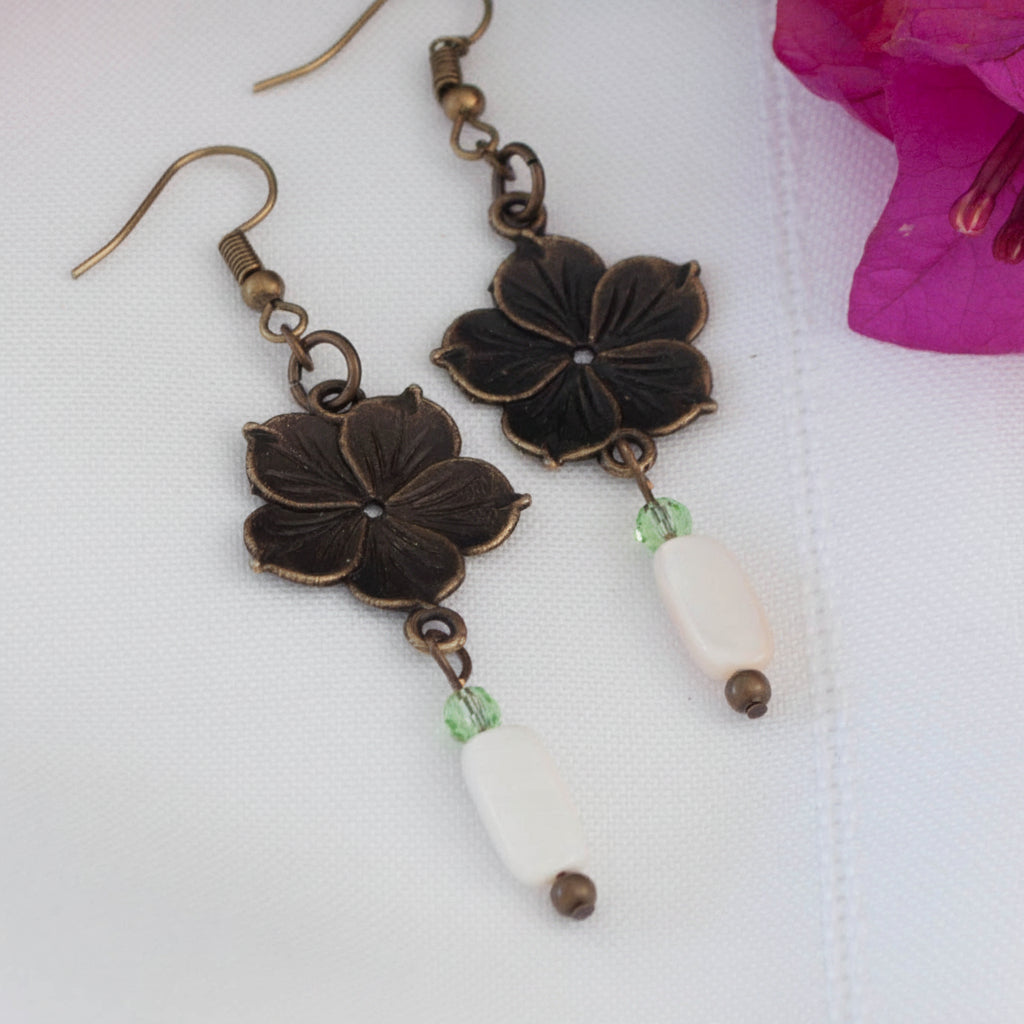 Pair of bronze flower charm earrings with mother of pearl drop and light green crystal bead, displayed on white surface with pink bougainvillea