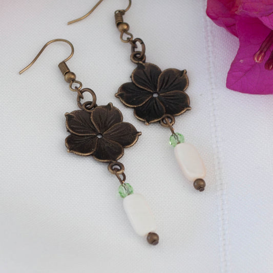 Pair of bronze flower charm earrings with mother of pearl drop and light green crystal bead, displayed on white surface with pink bougainvillea