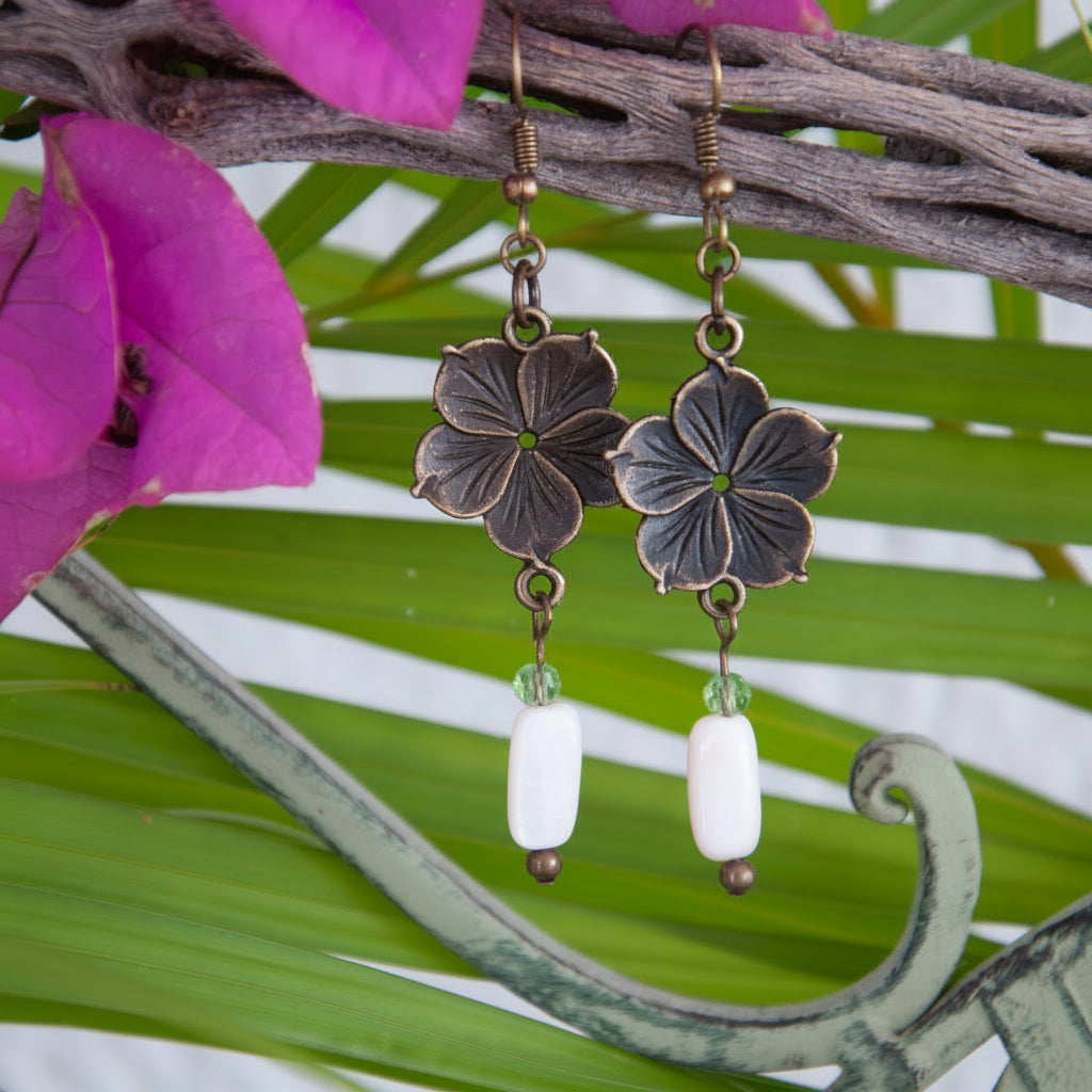 Bronze flower charm earrings with mother of pearl drop and light green crystal accent, hanging from driftwood with tropical palm leaves and bougainvillea
