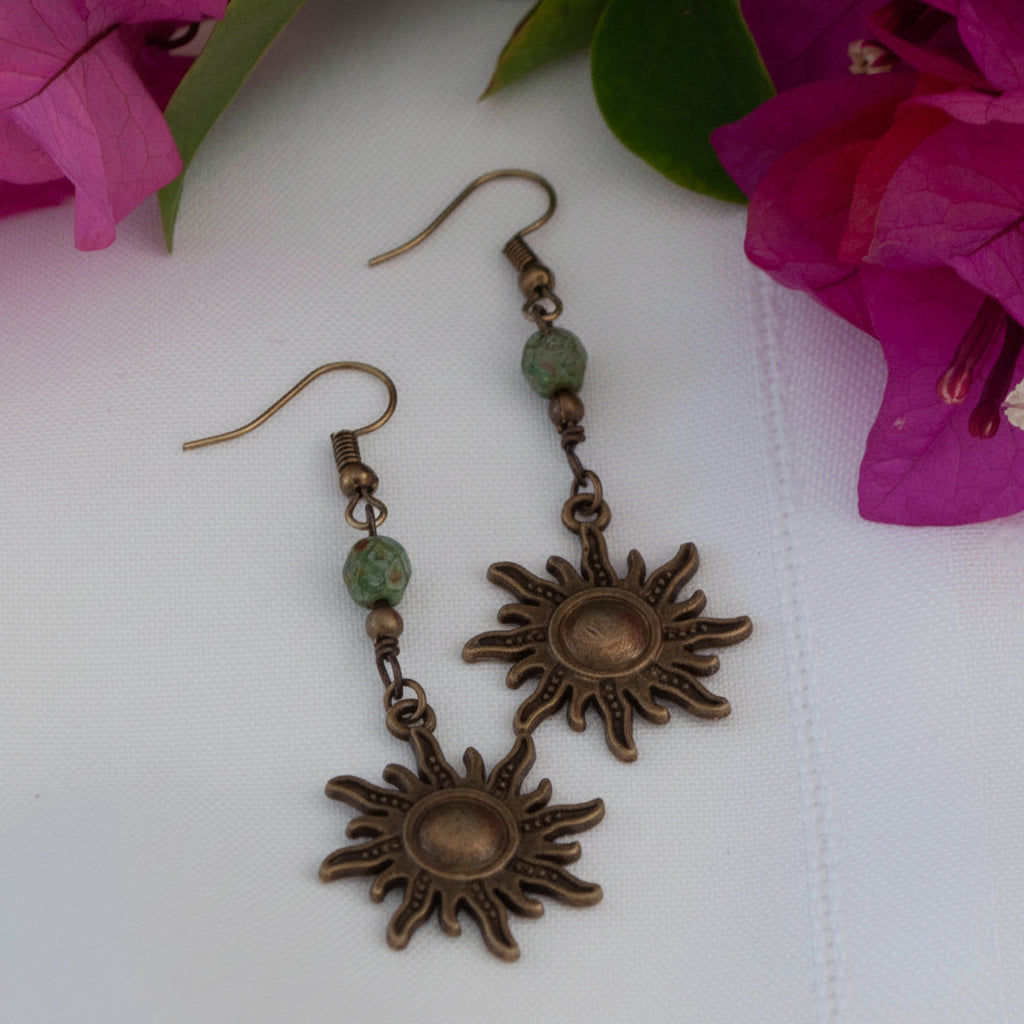 Pair of bronze sunburst charm earrings with green Picasso Czech beads and bronze accents, displayed on white surface with pink bougainvillea
