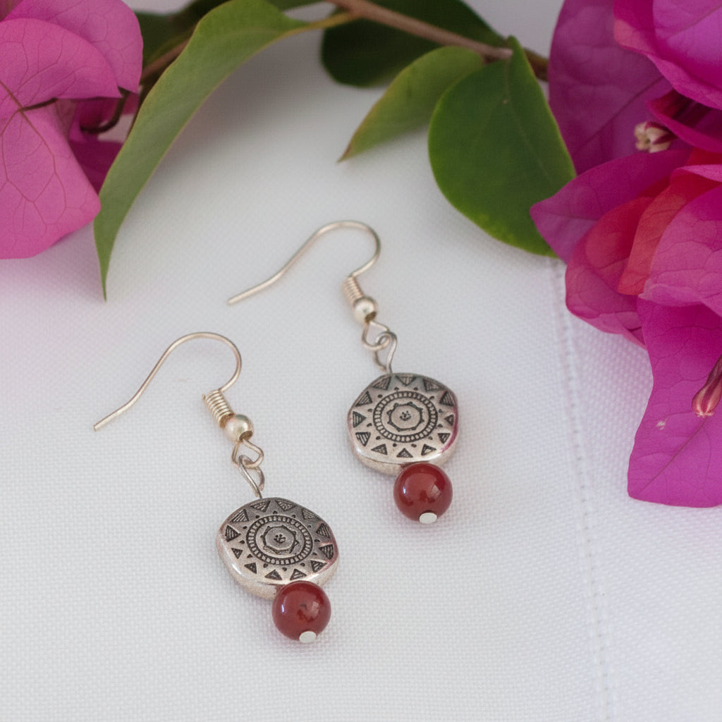 Handmade carnelian gemstone drop earrings with an Aztec-inspired silver-tone pewter disc bead and silver-tone fish hook wires, displayed on a white surface with magenta bougainvillea flowers.