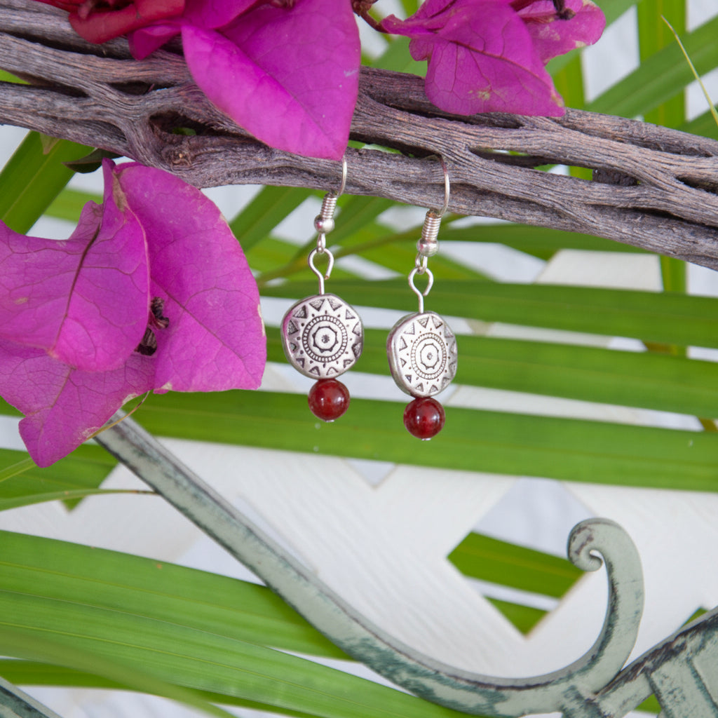 Handmade carnelian gemstone drop earrings with an Aztec-inspired silver-tone pewter disc bead and silver-tone fish hook wires, hanging from a driftwood branch with tropical palm leaves and magenta flowers.
