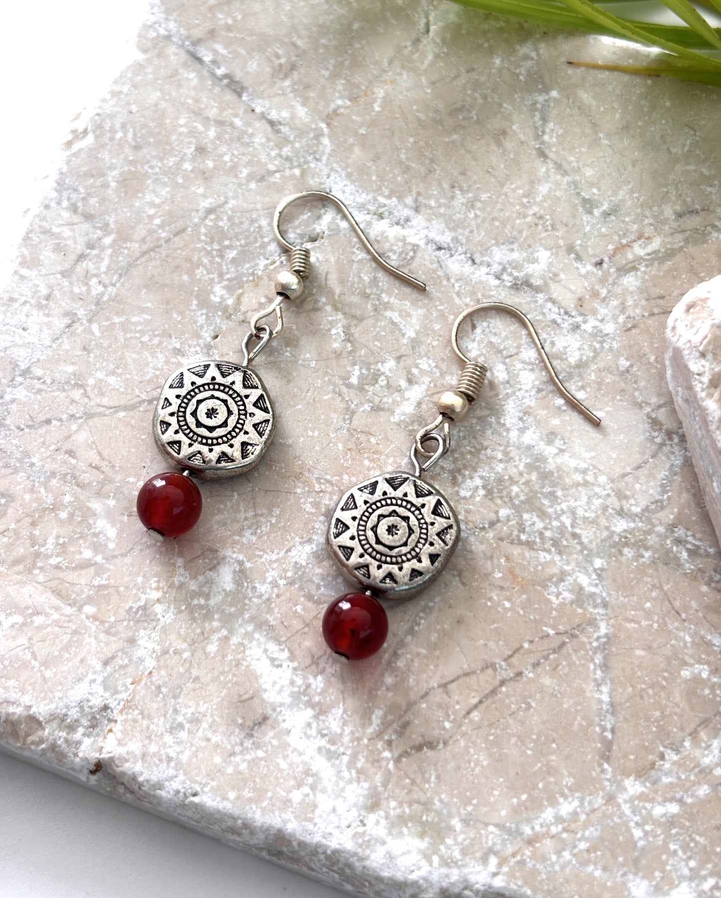 Pair of handmade carnelian gemstone drop earrings with Aztec-inspired silver-tone pewter disc beads and silver-tone fish hook wires, laid flat on a natural stone surface.