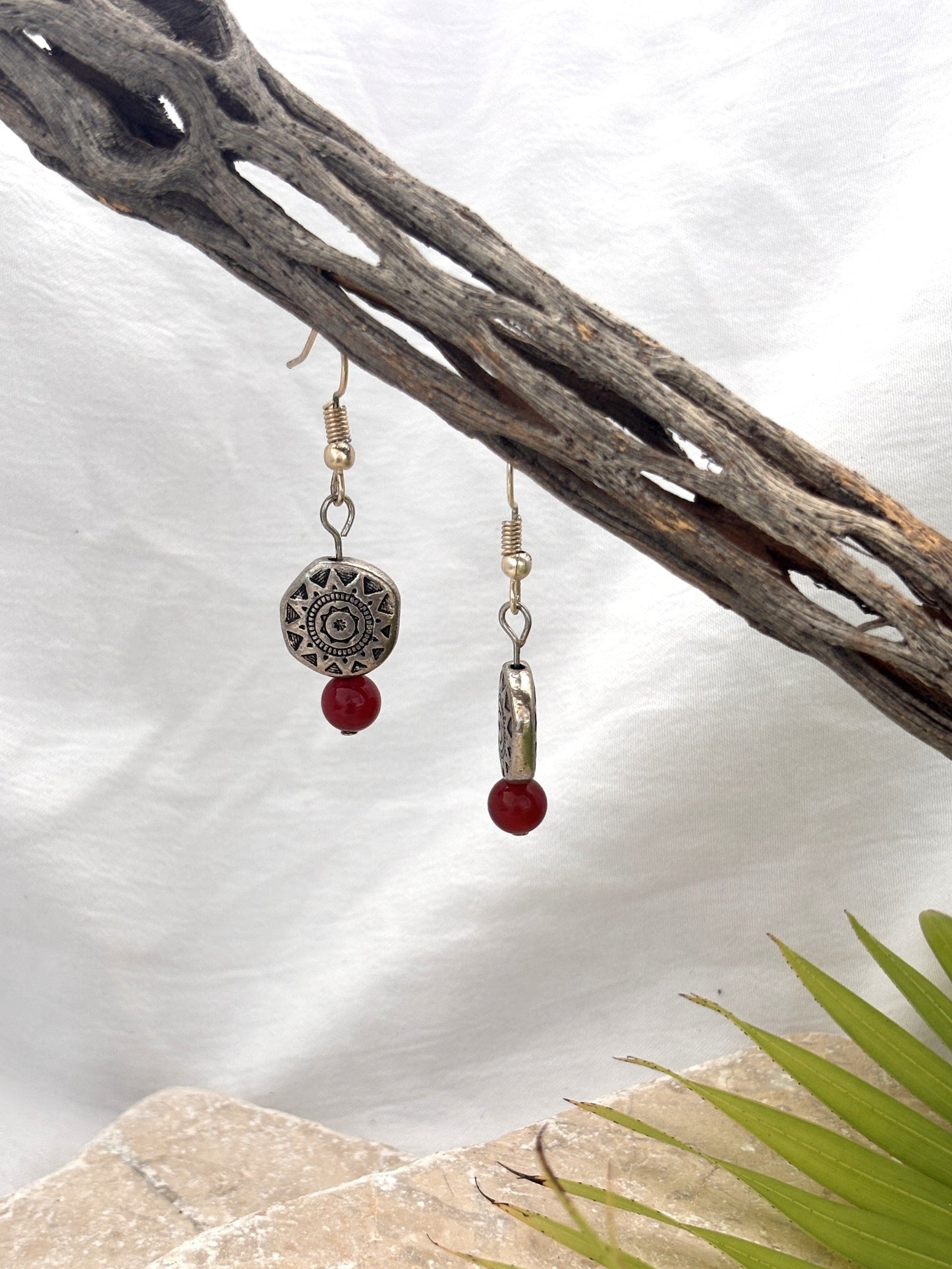 Handmade carnelian gemstone drop earrings with Aztec-inspired silver-tone pewter disc beads hanging from a driftwood branch with a stone and palm leaf backdrop