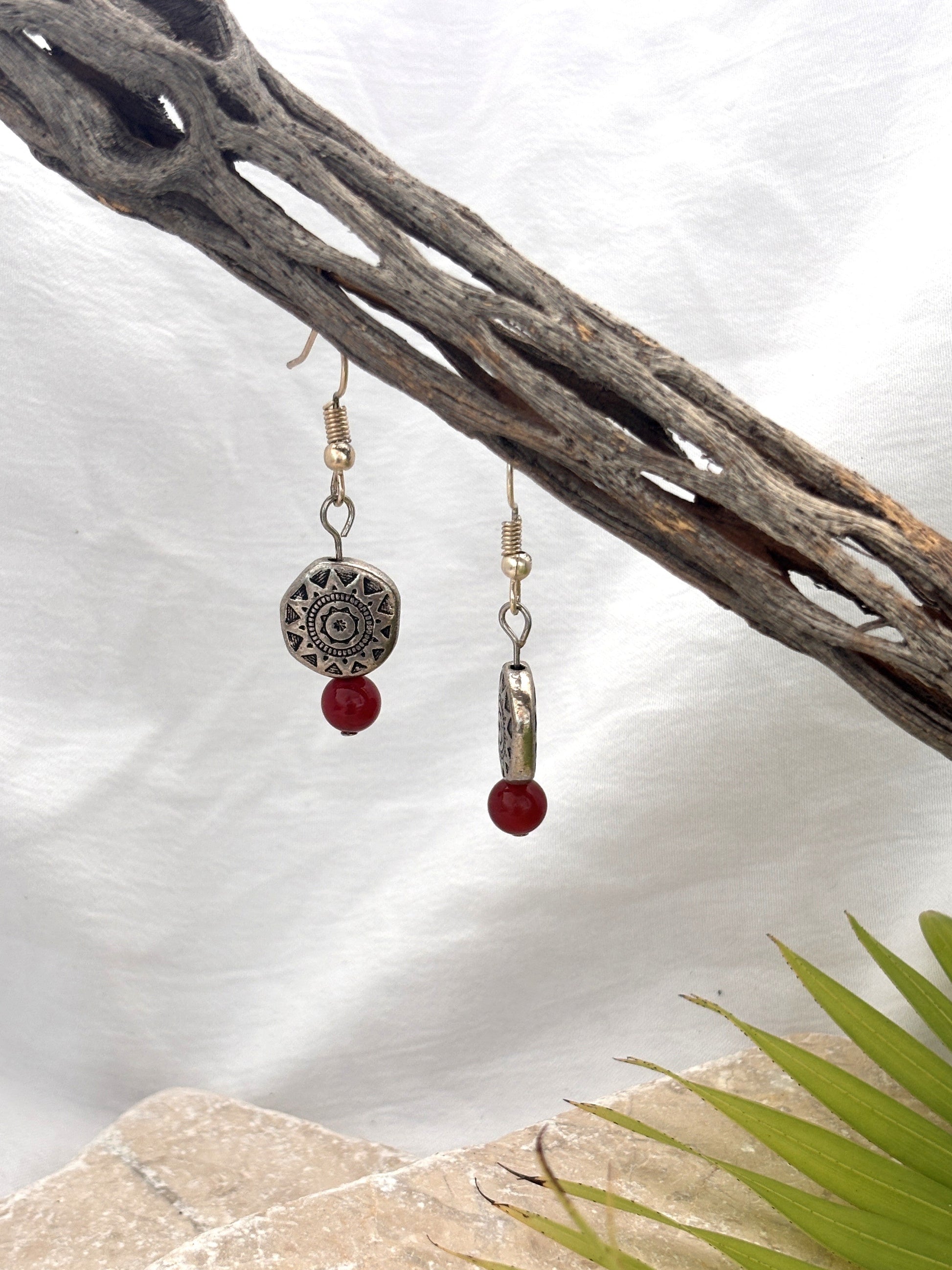 Handmade carnelian gemstone drop earrings with Aztec-inspired silver-tone pewter disc beads hanging from a driftwood branch with a stone and palm leaf backdrop