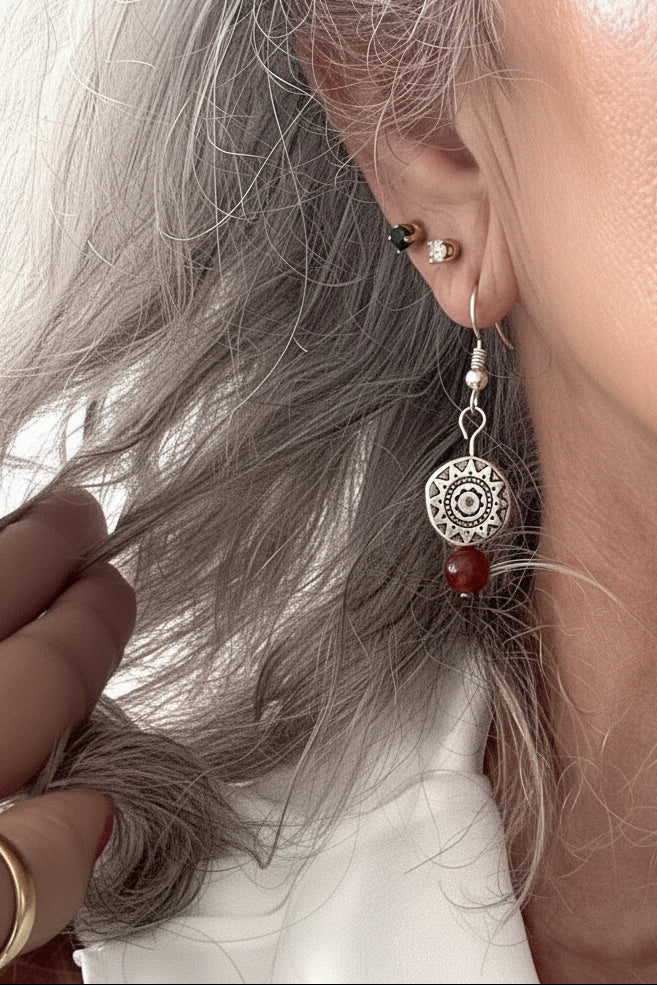 Close-up of a handmade carnelian gemstone drop earring with an Aztec-inspired silver-tone pewter disc bead on a silver-tone fish hook wire, worn on a model's ear.