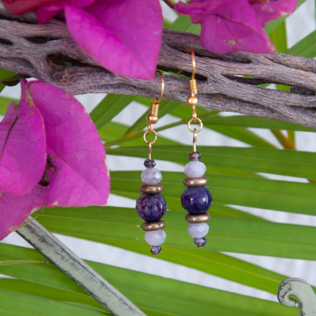 Handmade beaded drop earrings featuring an amethyst gemstone bead, bronze disc beads, and iridescent pink-and-purple faceted crystals on gold-tone fish hook wires, hanging from a driftwood branch with tropical palm leaves and magenta bougainvillea flowers.