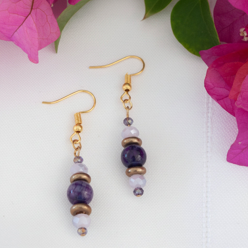 Handmade beaded drop earrings featuring an amethyst gemstone bead, bronze disc beads, and iridescent pink-and-purple faceted crystals on gold-tone fish hook wires, displayed on a white surface with magenta bougainvillea flowers.