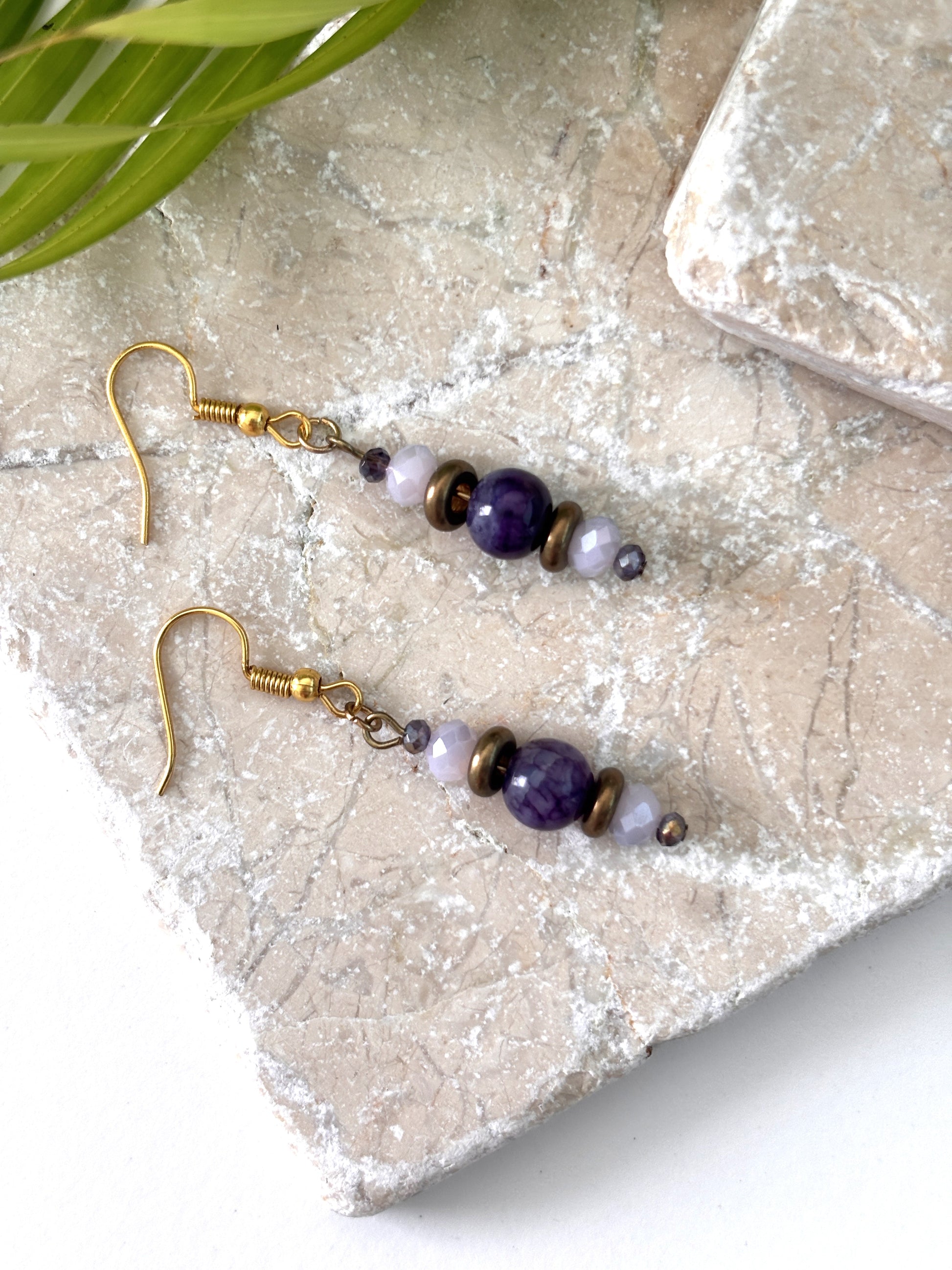 Handmade beaded drop earrings with an amethyst gemstone bead, bronze disc beads, and iridescent faceted crystals on gold-tone fish hook wires, laid flat on a natural stone surface with a tropical palm leaf.