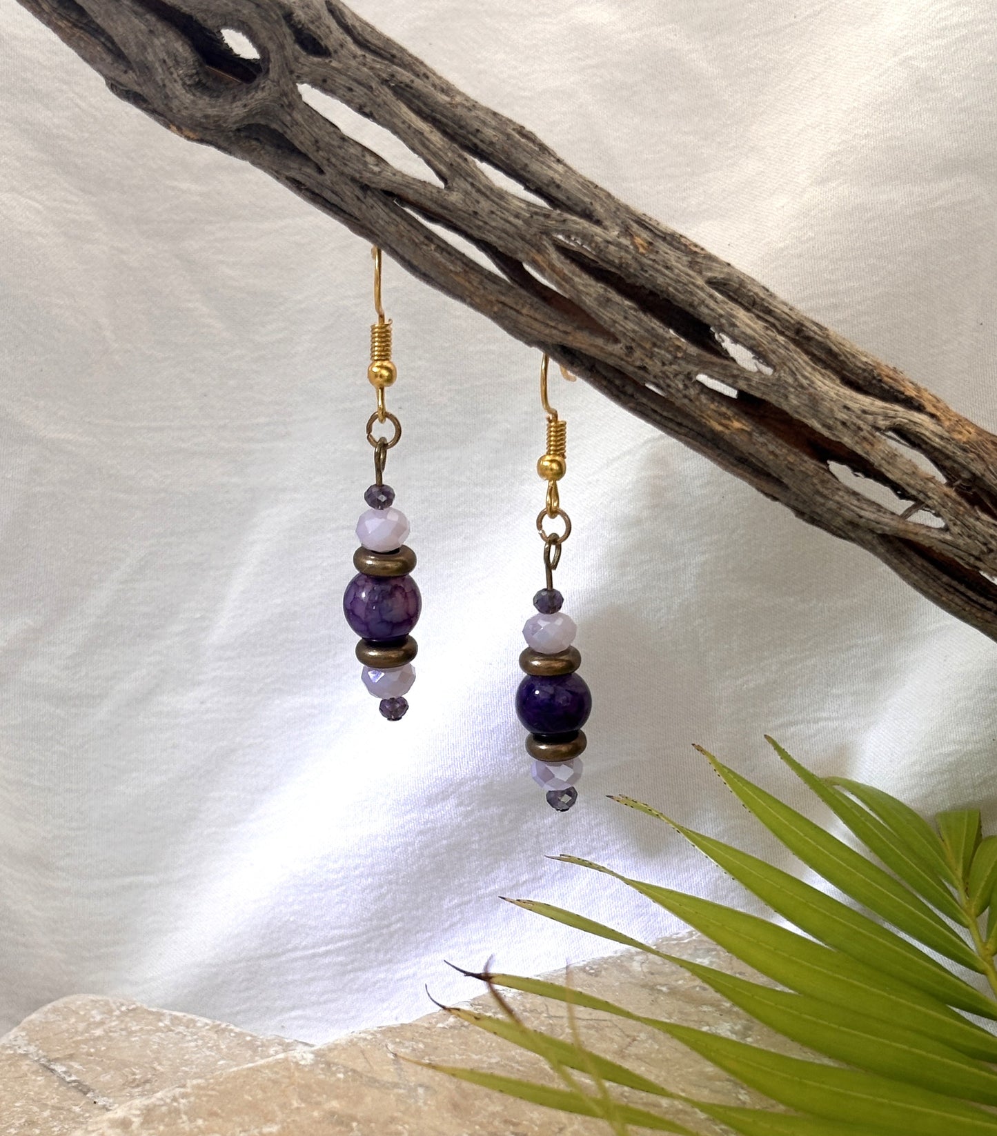 Handmade beaded drop earrings with an amethyst gemstone bead, bronze disc beads, and iridescent faceted crystals hanging from a driftwood branch with a stone and palm leaf backdrop.